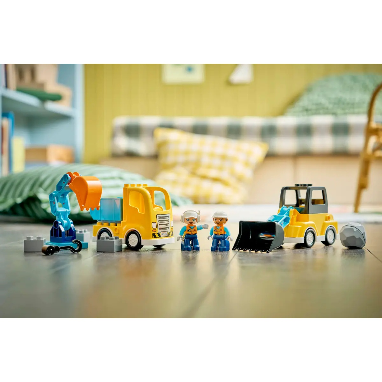 Une scène de construction avec des camions jouets, une pelle et des ouvriers sur un sol en bois. L'arrière-plan comprend des oreillers et une chaise en bois.