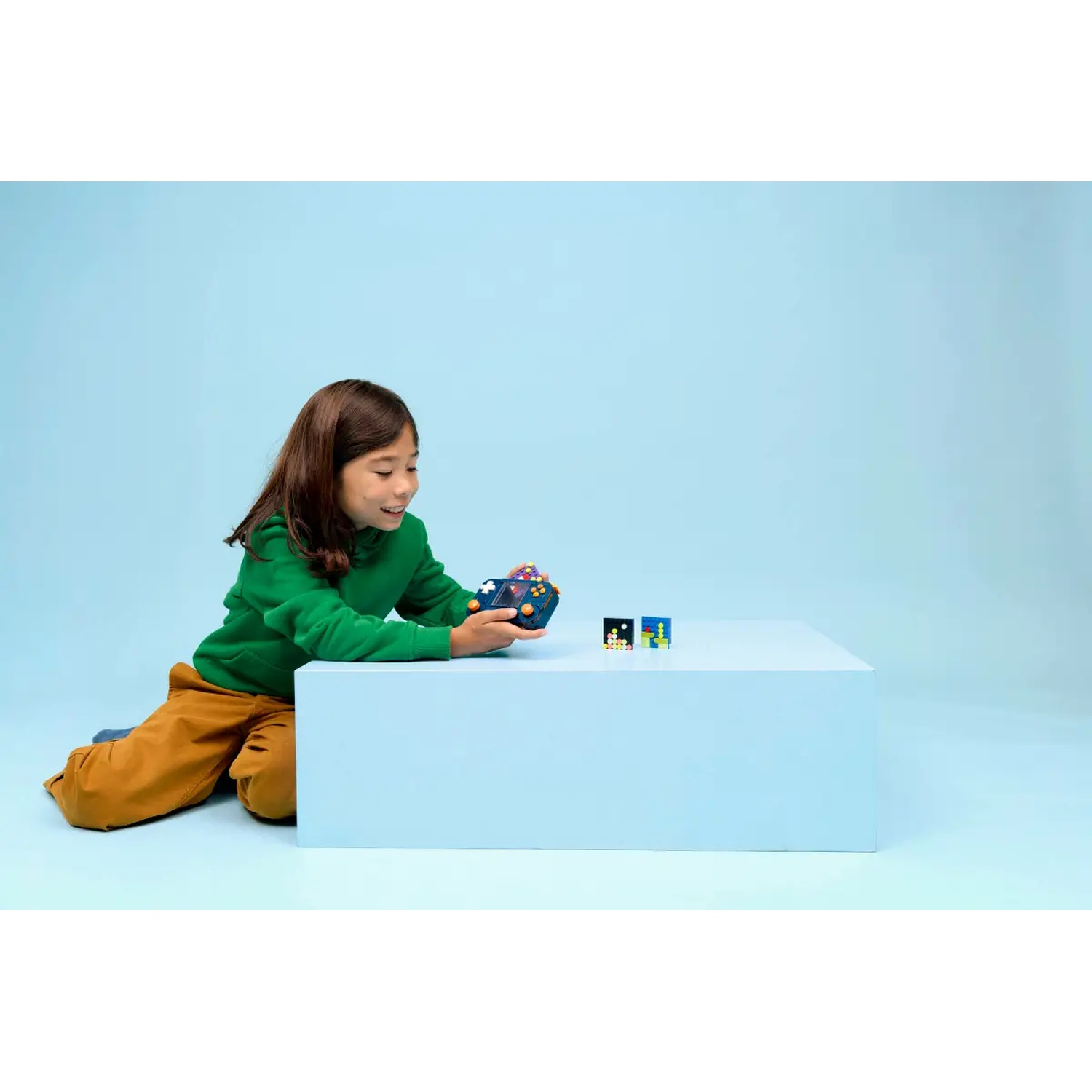 A girl in green top and brown pants is playing with a colorful toy. Two small toy cubes are on a light blue table.