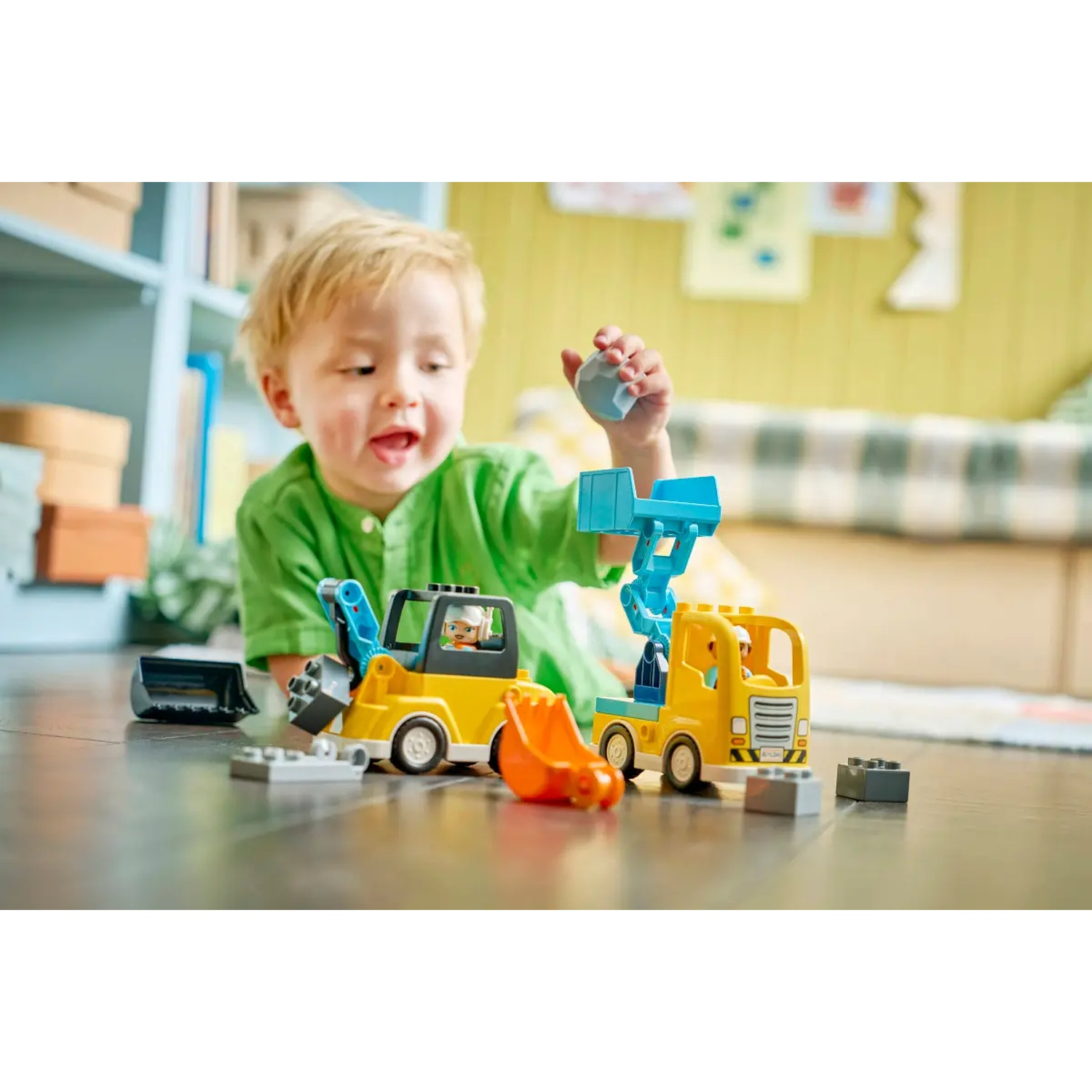 Un enfant joue avec des véhicules de chantier jouets sur un sol en bois. Une pelle et un camion jaunes sont devant l'enfant.