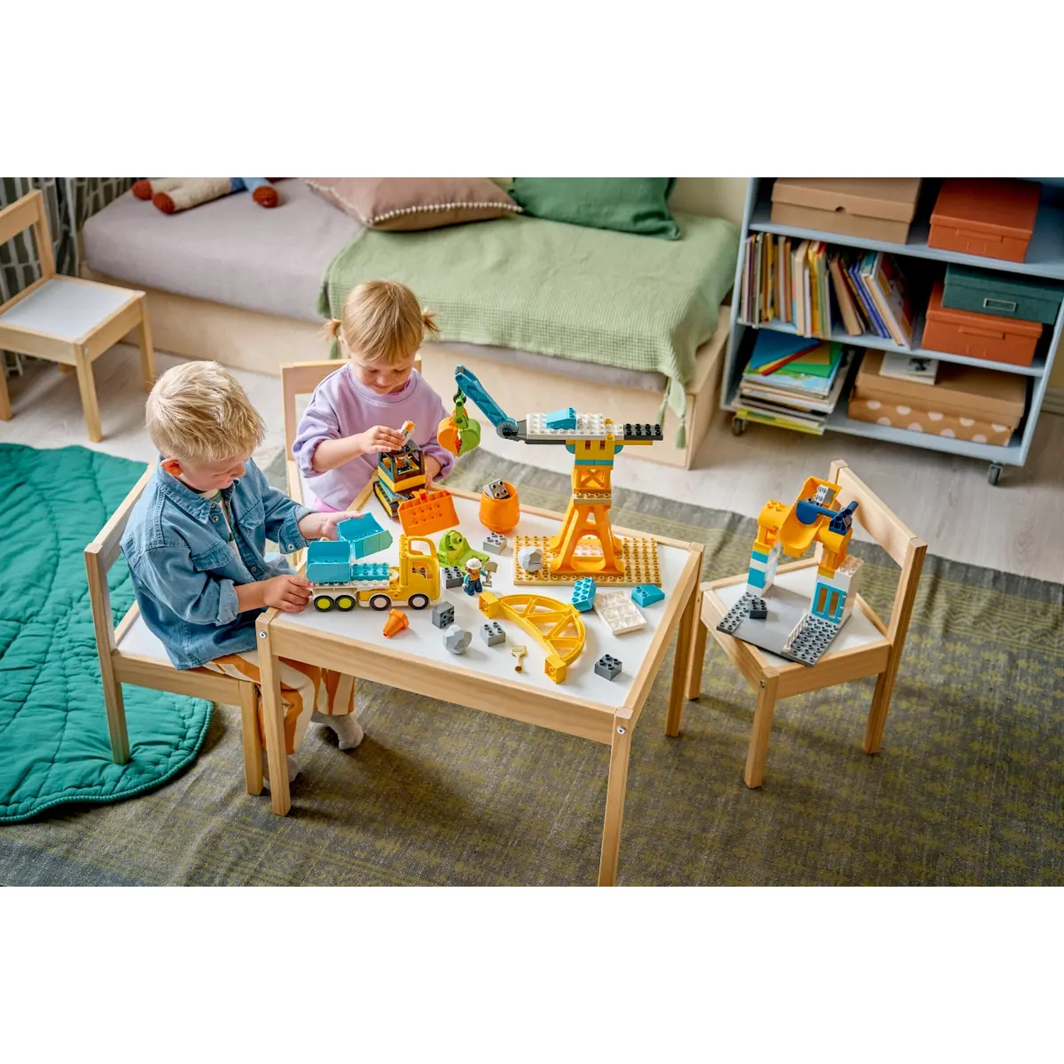 Deux enfants jouent avec des jouets de construction sur une table blanche. Une grue, un camion et une excavatrice sont présents. Fond : lit, étagères.