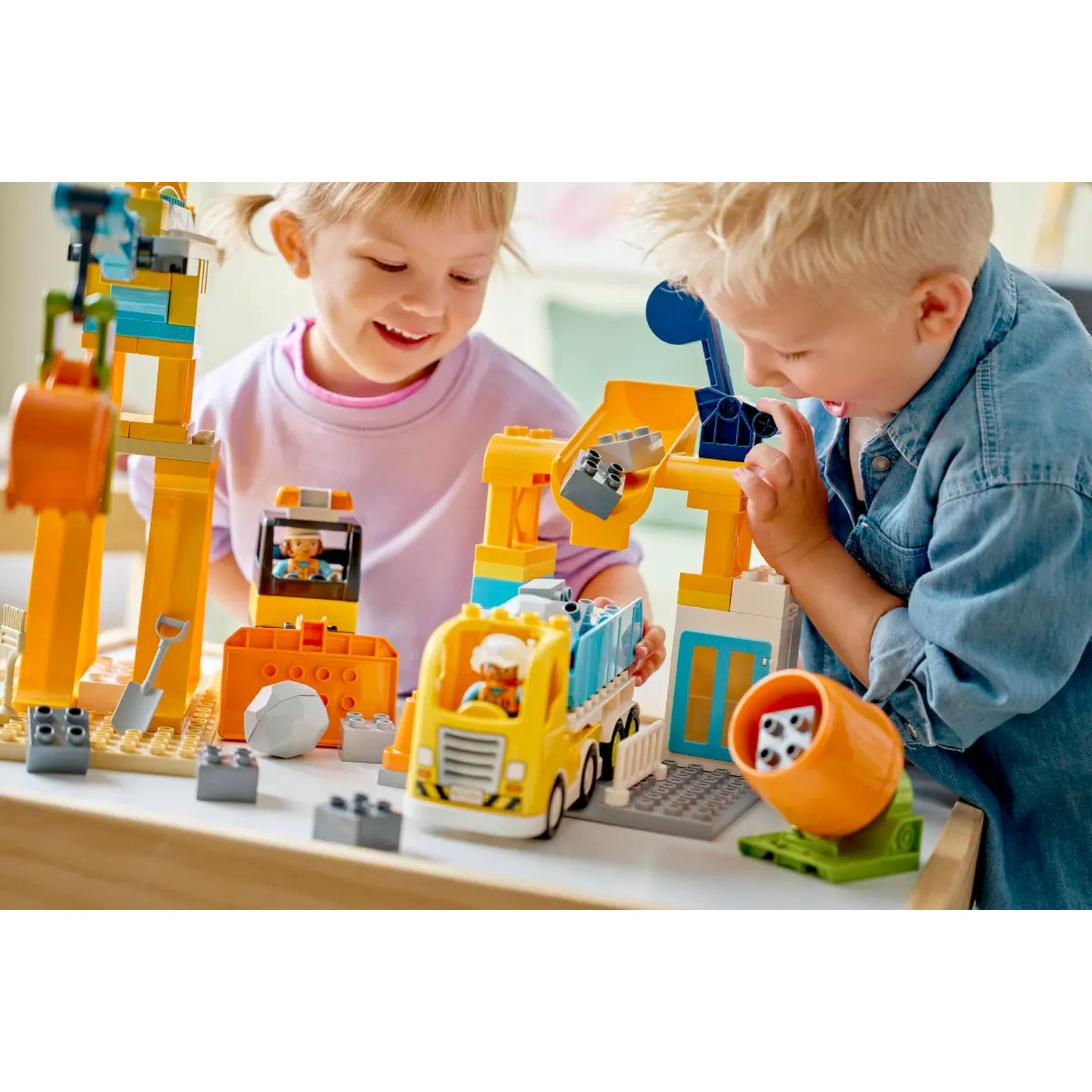 Deux enfants jouent avec un jeu de construction. Un camion jaune et une grue sont visibles. Les enfants sourient et sont concentrés sur leur jeu.