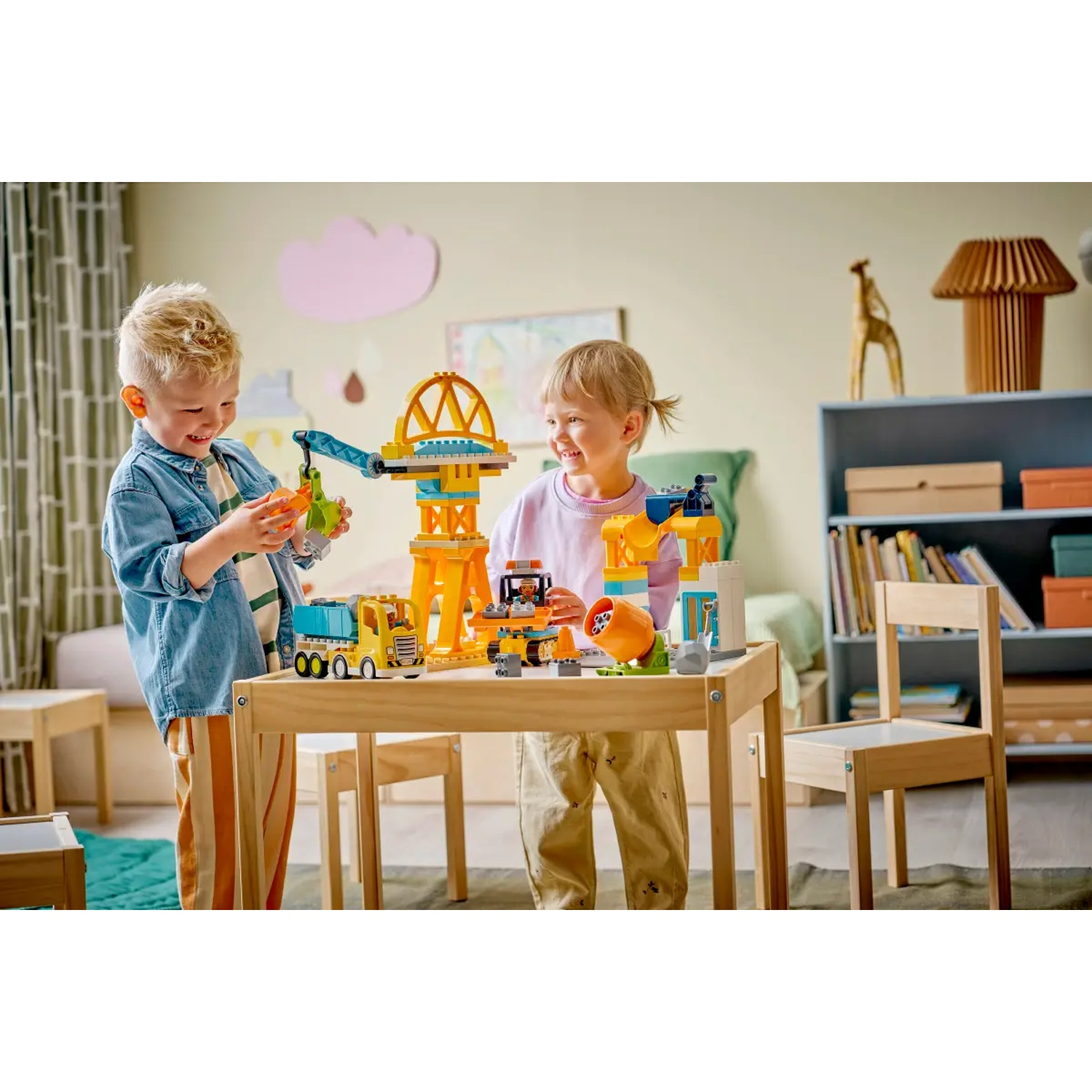 Deux enfants jouent avec un jeu de construction sur une table en bois. L'ensemble comprend une grue, des camions et d'autres éléments.