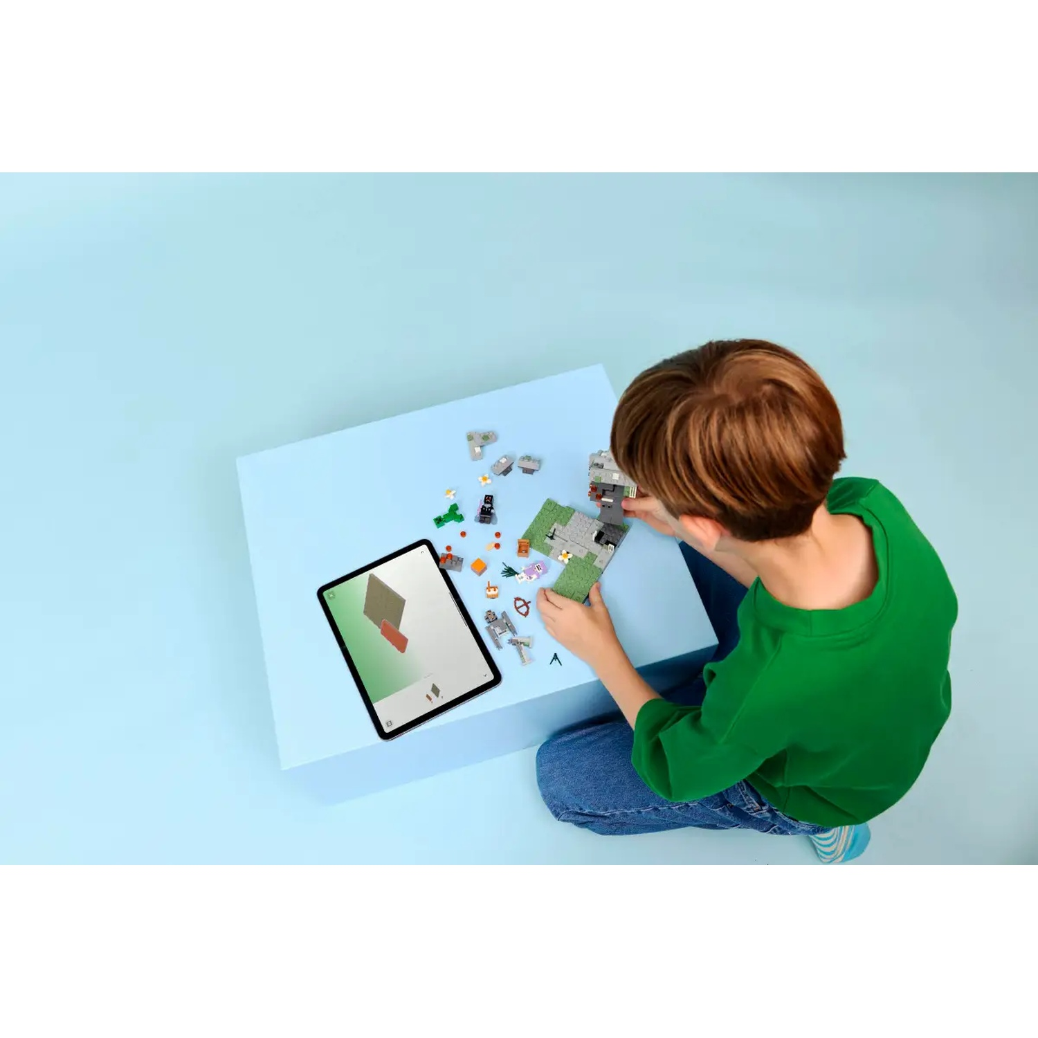 Une personne en chemise verte et jean assemble des blocs Lego avec une tablette sur une table bleue. De nombreux blocs sont dispersés.