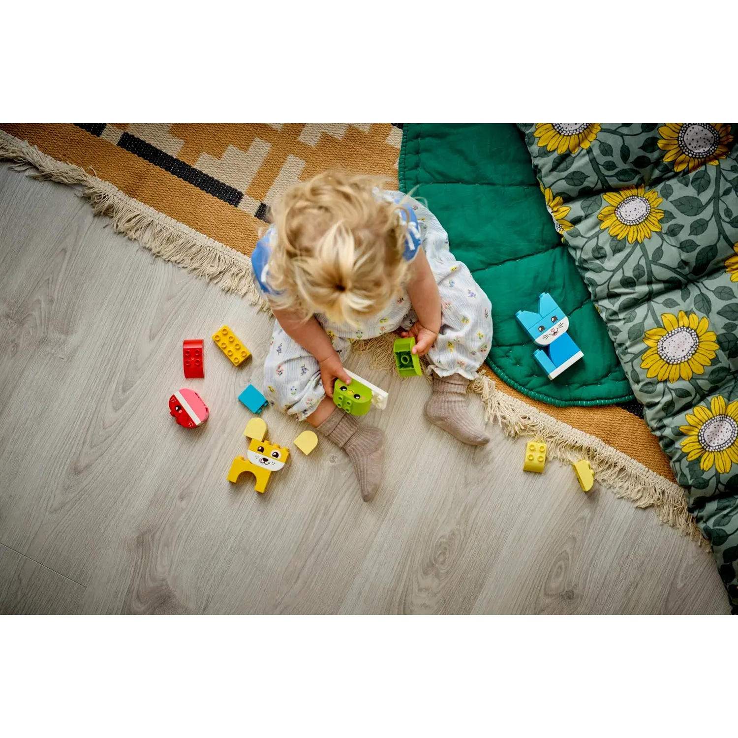 Un enfant joue avec des blocs de jouets colorés, un chien jaune et un lapin bleu sur le sol. Un tapis à motifs et un tapis vert sont à proximité.