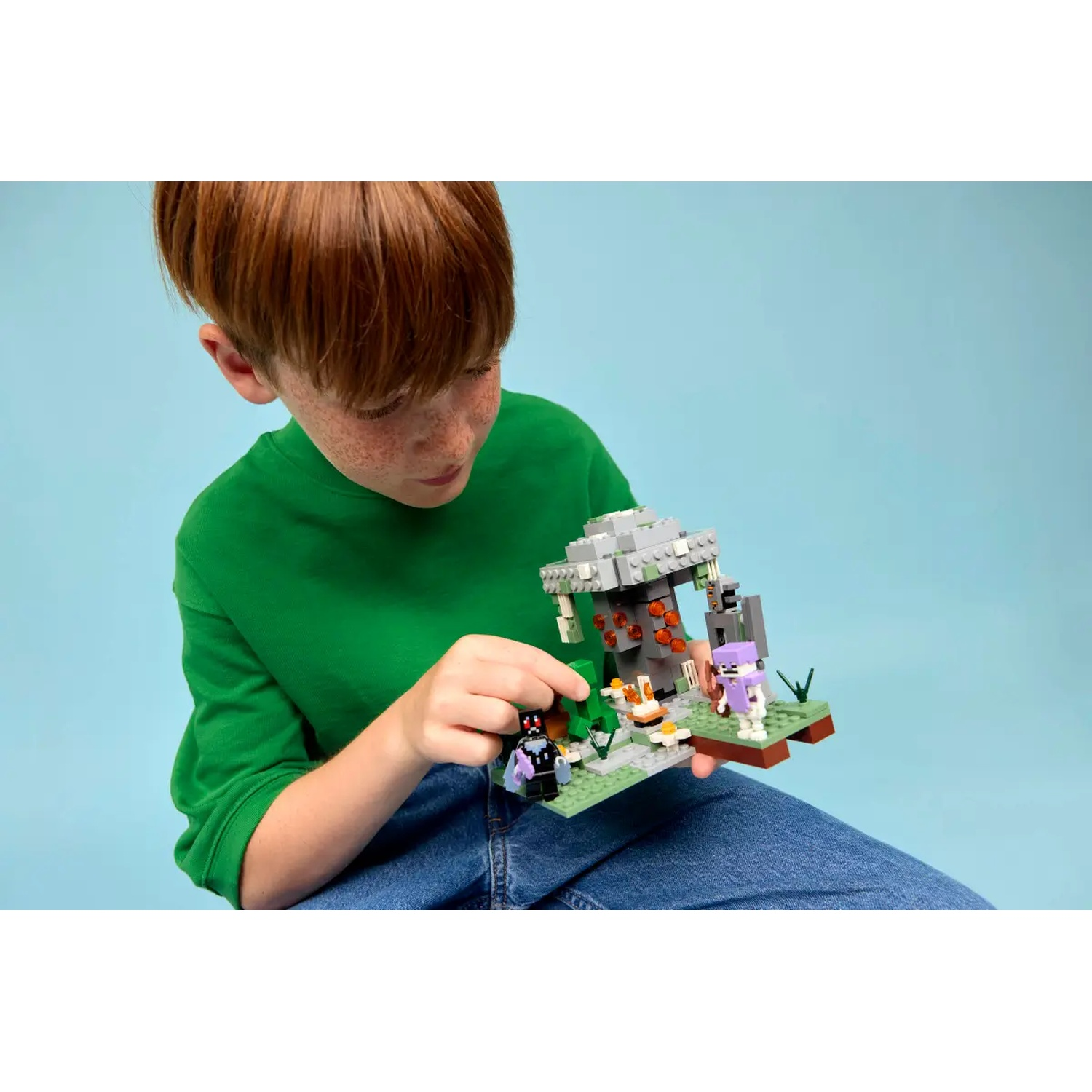 Un garçon aux cheveux roux et un t-shirt vert tient un jeu Lego Minecraft. L'ensemble représente des personnages et une scène de grotte. Fond bleu.