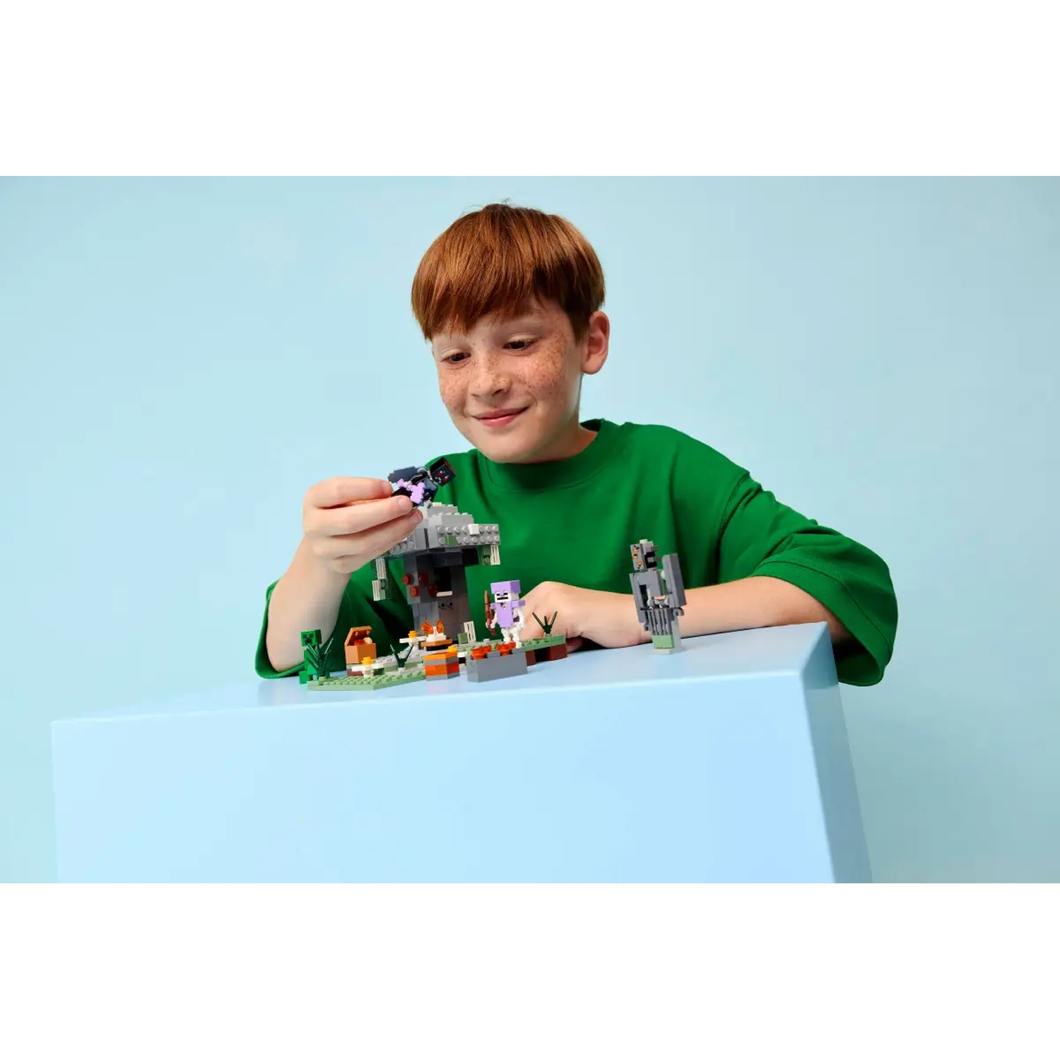Un garçon en chemise verte joue avec un jeu Lego Minecraft. L'ensemble est sur une table bleue. Le fond est bleu clair.