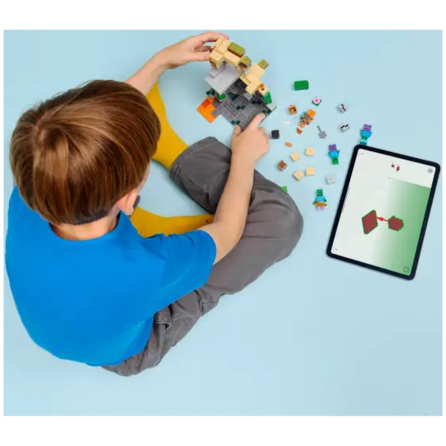 Un enfant construit avec des blocs Lego tout en regardant les instructions sur une tablette. Divers blocs et figurines sont éparpillés sur une surface bleue.