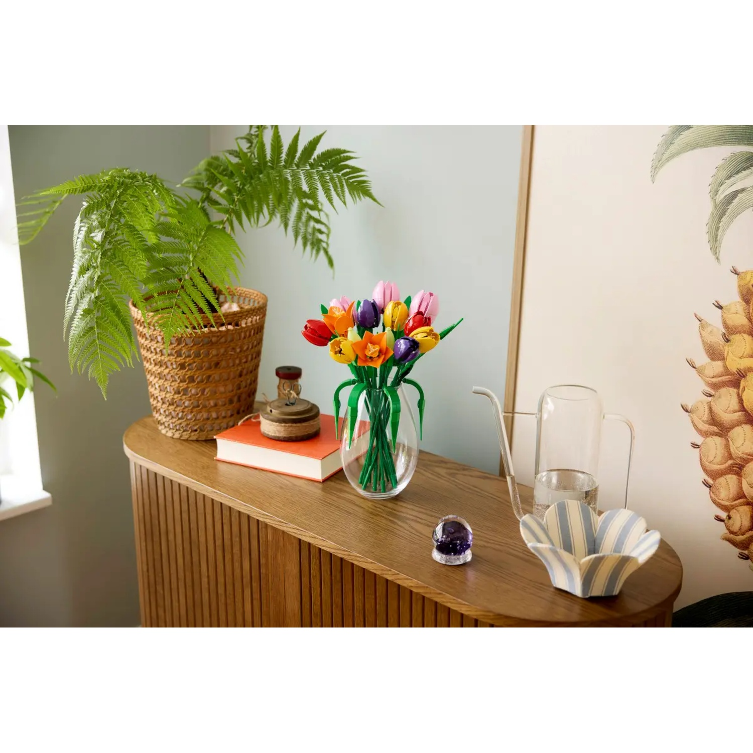 Een houten tafel toont een varen in een mand, kleurrijke tulpen in een vaas, een boek en decoratieve items. Achtergrond met ananaskunst.