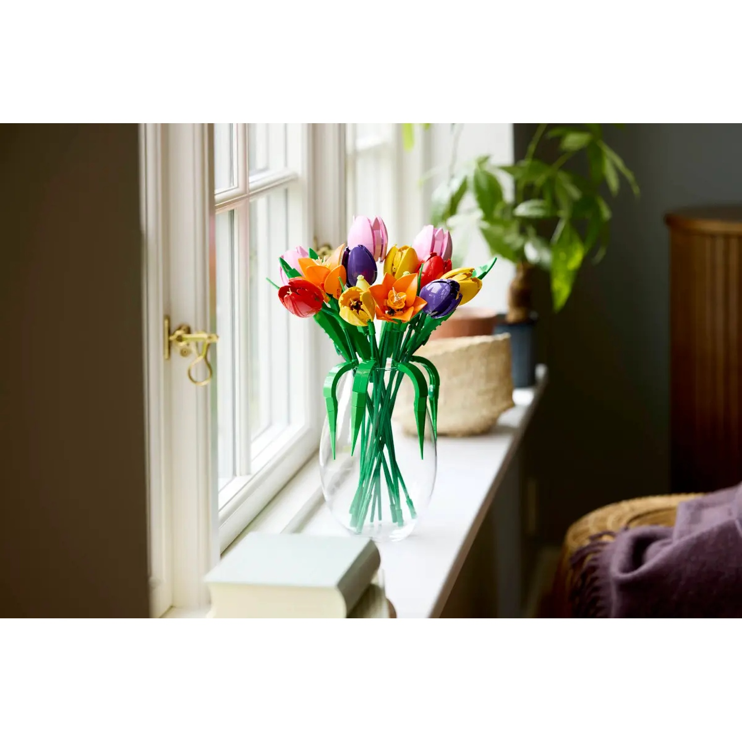 Kleurrijke Lego tulpen in een doorzichtige vaas op een vensterbank. Een boek, een mand en een plant staan ook op de vensterbank.
