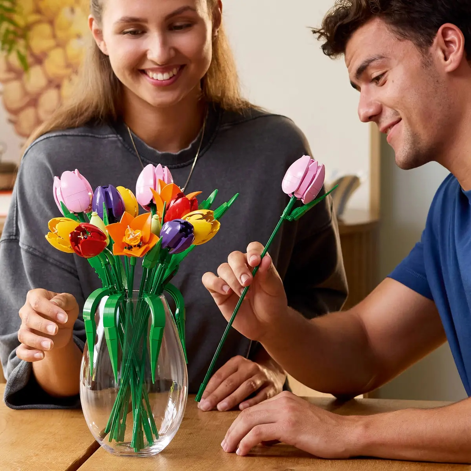 Een stel glimlacht en bewondert Lego-bloemen in een vaas. Een man houdt een enkele roze tulp vast. Kleuren: roze, oranje, geel en paars.