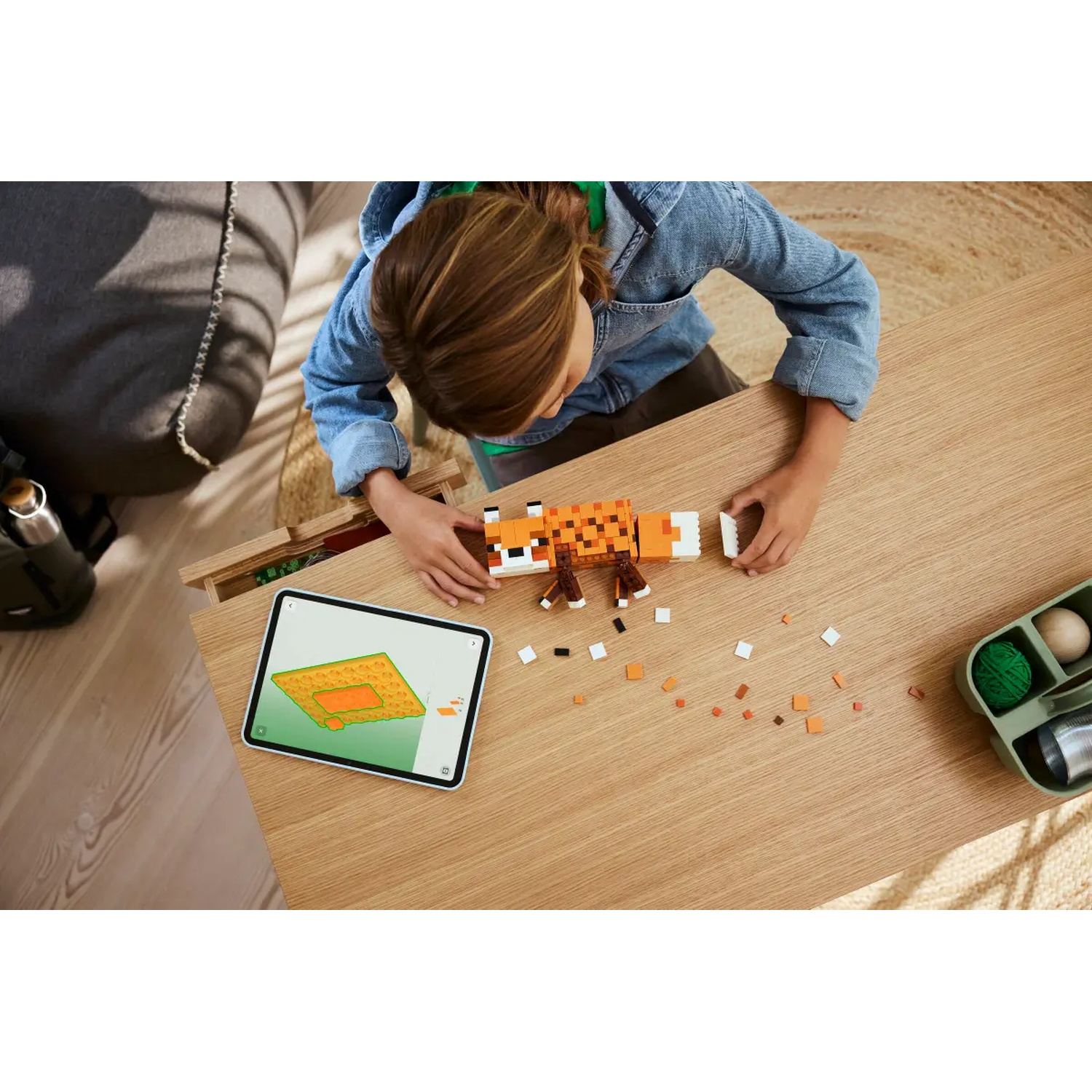 Un enfant construit un jouet renard pixélisé sur une table en bois. Une tablette affiche des instructions. Des blocs éparpillés et un récipient sont à proximité.