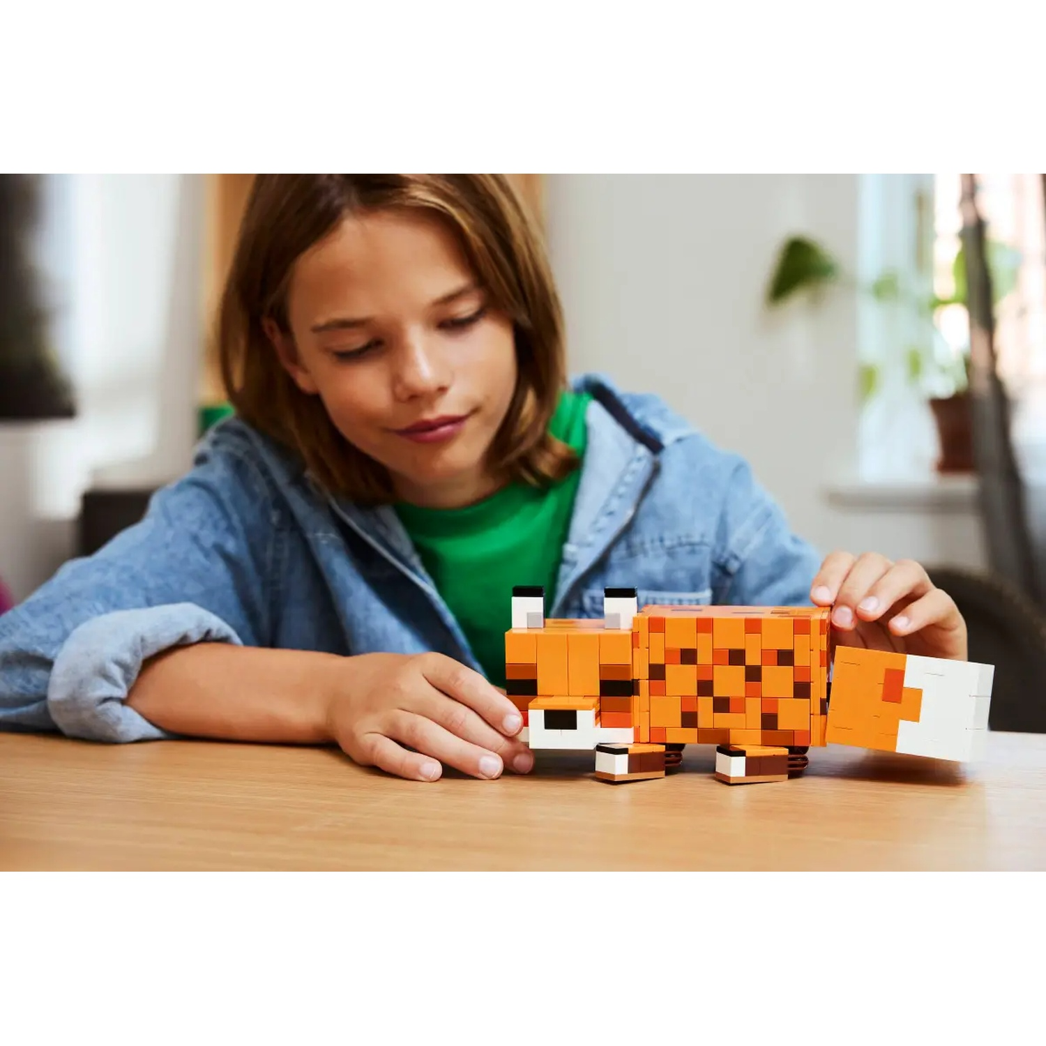 Un enfant joue avec un renard Minecraft construit avec des blocs sur une table en bois. L'enfant porte une veste en jean.