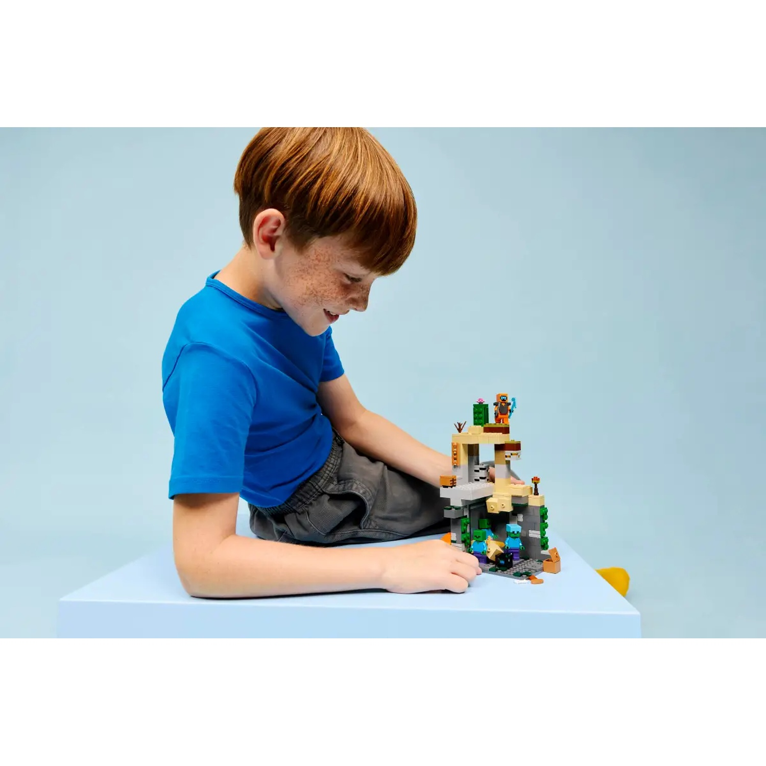 Un garçon roux en chemise bleue regarde un jeu Lego Minecraft sur une surface bleue. Le fond est également bleu.