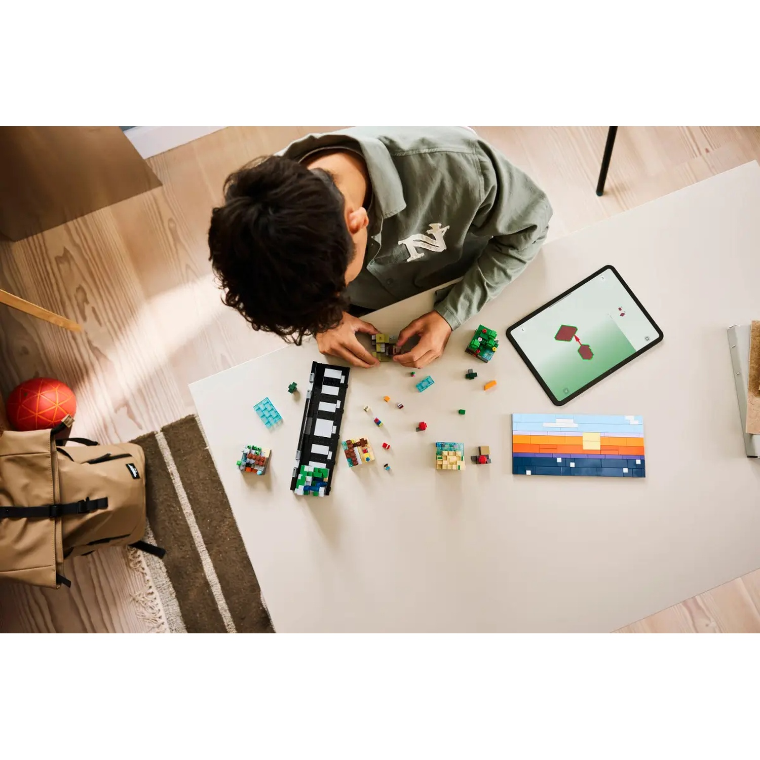 Une personne construit avec des blocs colorés sur une table blanche. Un iPad montre un guide de construction. Un sac et une balle à gauche.