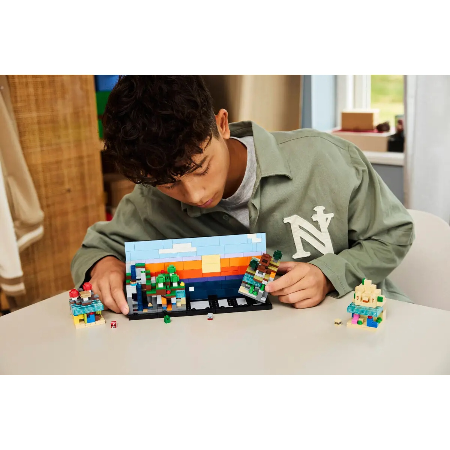 Une personne construit un diorama Lego. Le diorama représente un paysage avec ciel bleu, arbres et coucher de soleil. Il y a d'autres structures Lego.