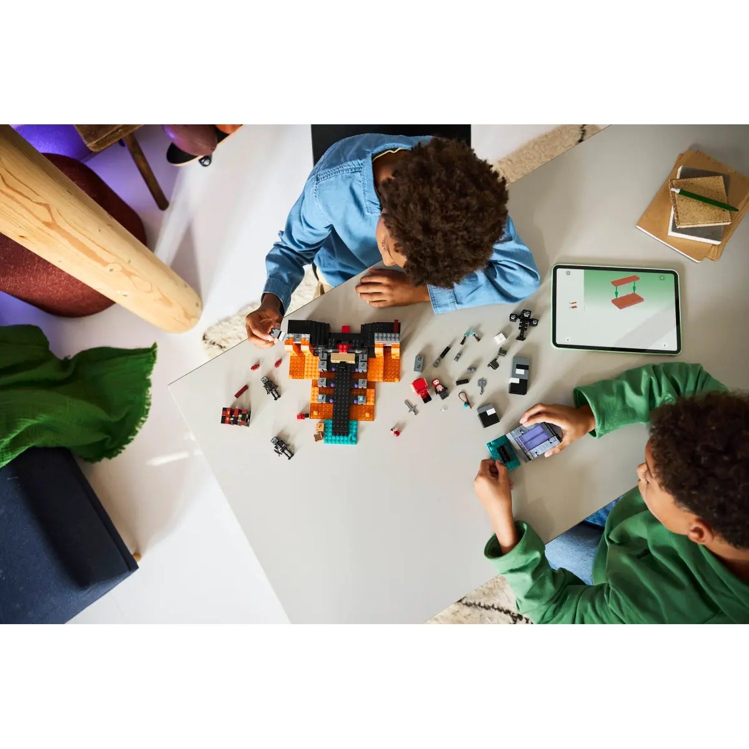 Deux enfants construisent avec des Lego sur une table blanche. Une tablette et des pièces Lego sont dispersées. Une batte de baseball et un tissu vert sont visibles.