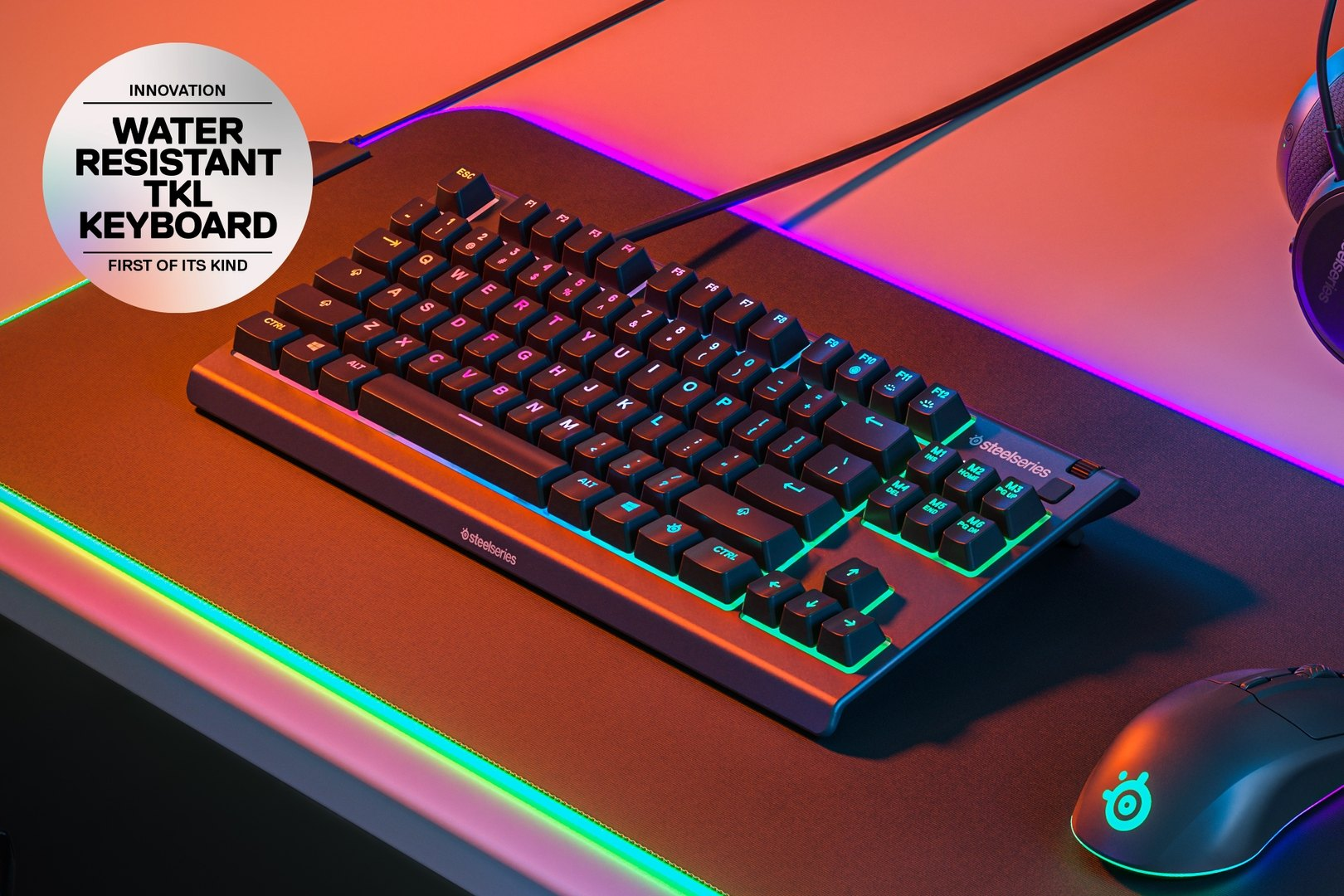 Eine wasserdichte TKL-Tastatur von SteelSeries mit leuchtenden Tasten liegt auf einer Matte neben einer Maus, ein Headset ist im Hintergrund sichtbar.
