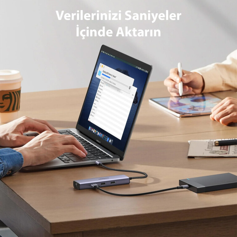 Bir kişi, dosya aktarımı yapan bir hub'a bağlı bir dizüstü bilgisayar kullanıyor. Ahşap masada bir tablet ve kahve fincanı da var.