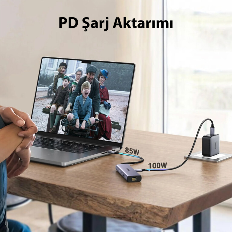 Ekranında film sahnesi olan, şarj cihazına ve bir hub'a bağlı dizüstü bilgisayar. Ekranda metin: PD Şarj Aktarımı. Kablolar 85W ve 100W gösteriyor.