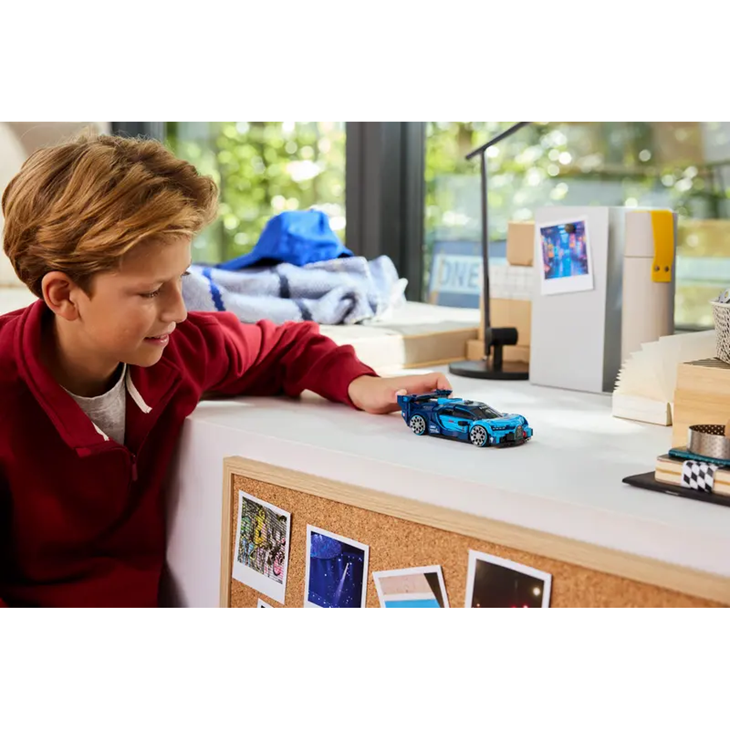Een jongen bekijkt een blauwe speelgoedauto op een bureau. Achter hem is een raam. Er zijn foto's op een bord gespeld.