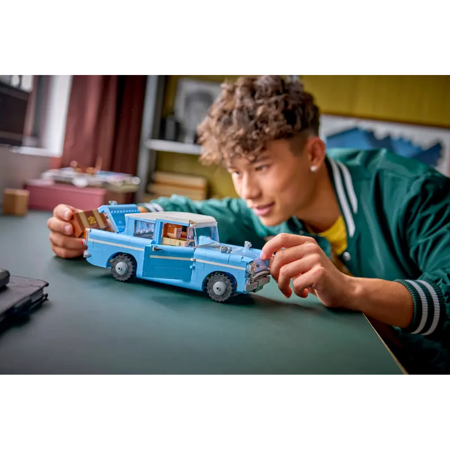 A young man examines a light blue Lego car. The car is on a table. He wears a green jacket, and a yellow shirt.