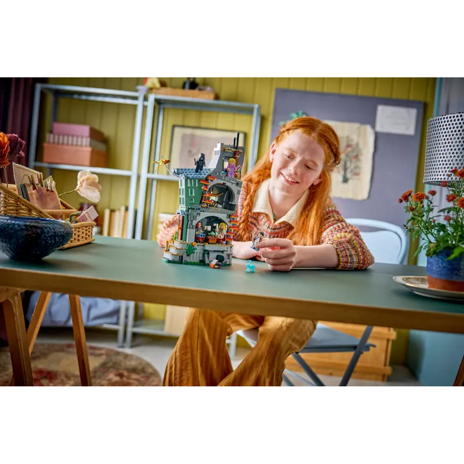 A girl with red hair smiles, playing with Lego. A Lego building sits on a green table. Other objects are visible in the background.