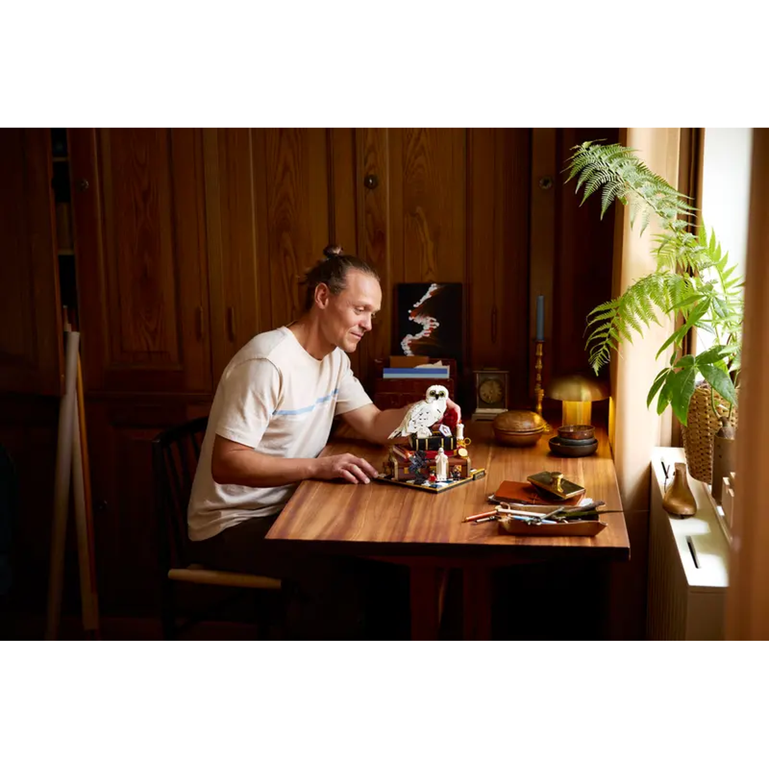 A man sits at a wooden table, looking at a white owl figurine. A window with plants is on the right. A painting is on the left.