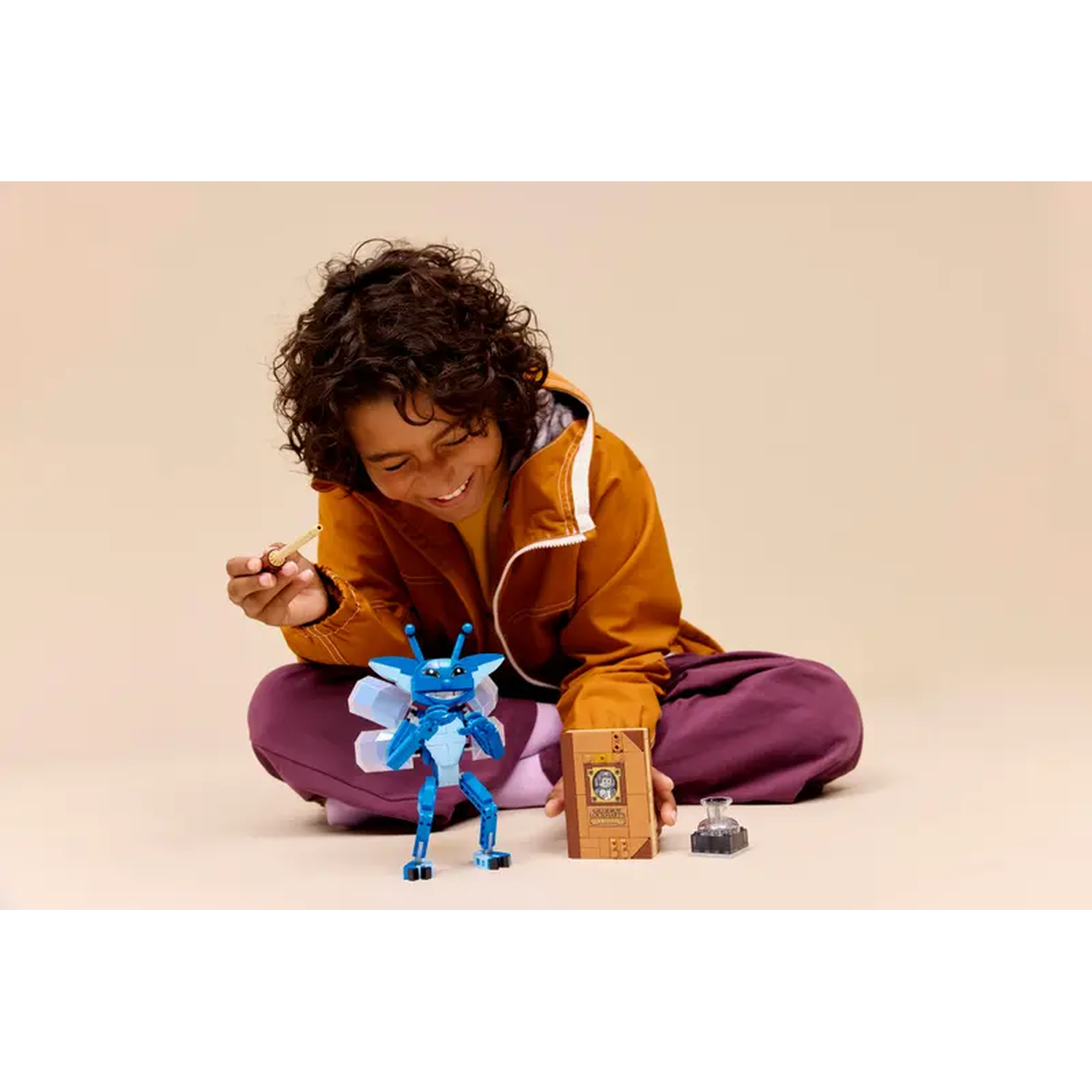 A smiling child holds a stick, surrounded by a blue insect toy, a book, and a small inkwell. They wear a brown jacket and purple pants.