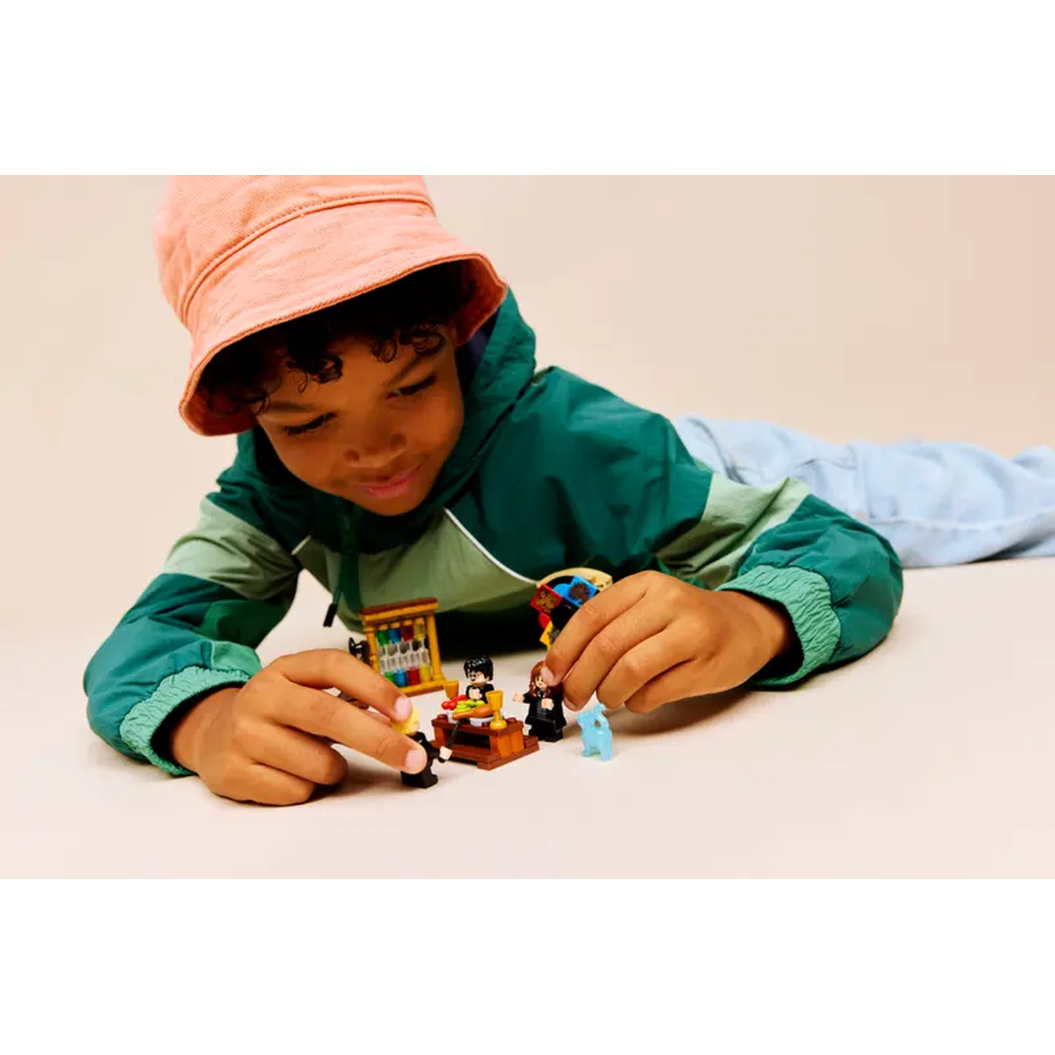 A child in a green jacket and pink hat plays with Lego figures. The child is focused on the figures arranged on a plain surface.