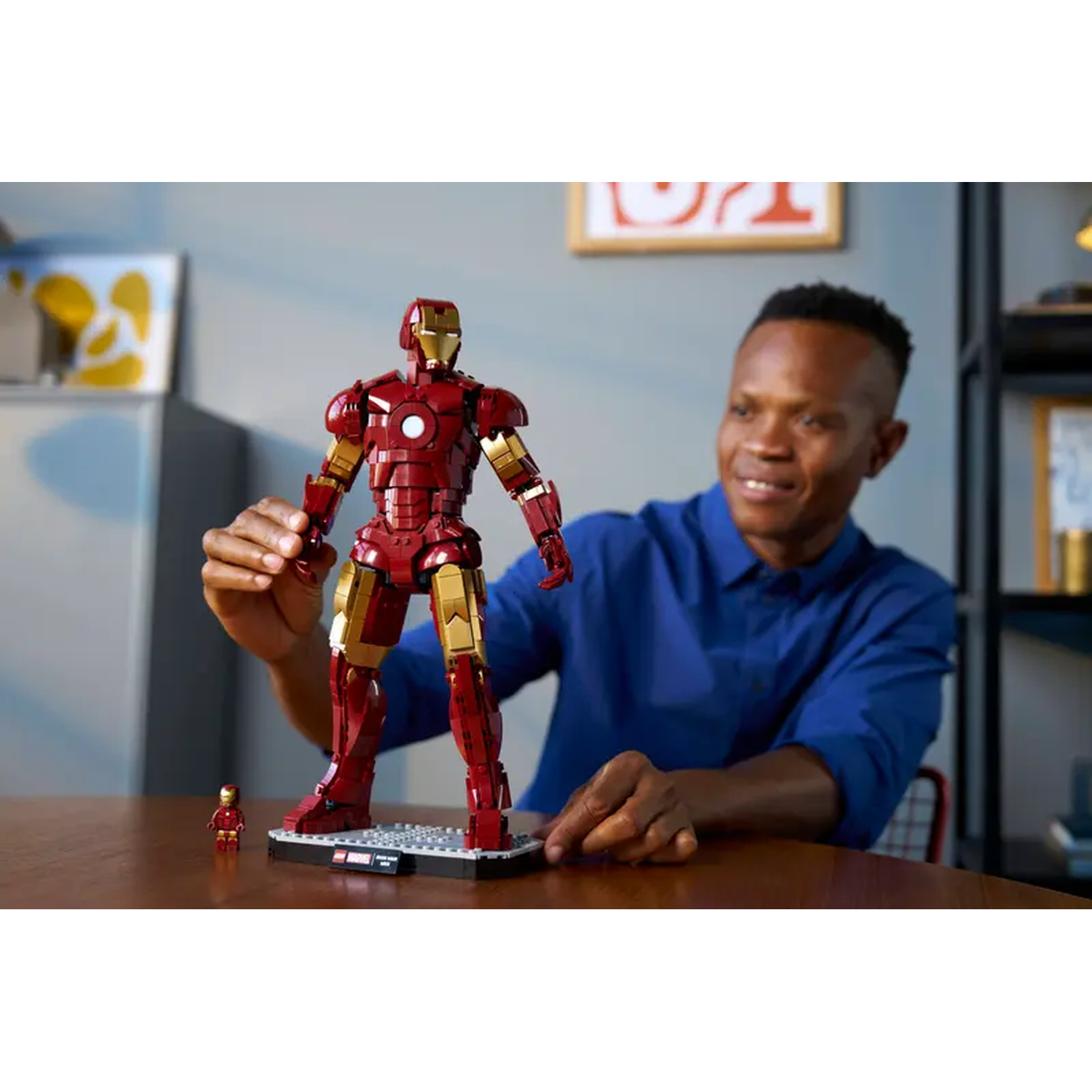A man smiles, looking at a large Lego Iron Man. Smaller Iron Man figurine in front. Red and gold armor.