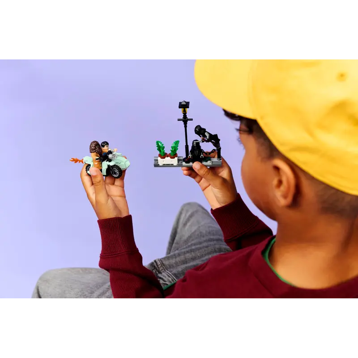 A child holds Lego sets: a motorcycle with figures and a street scene with a lamppost and figures. Purple background.