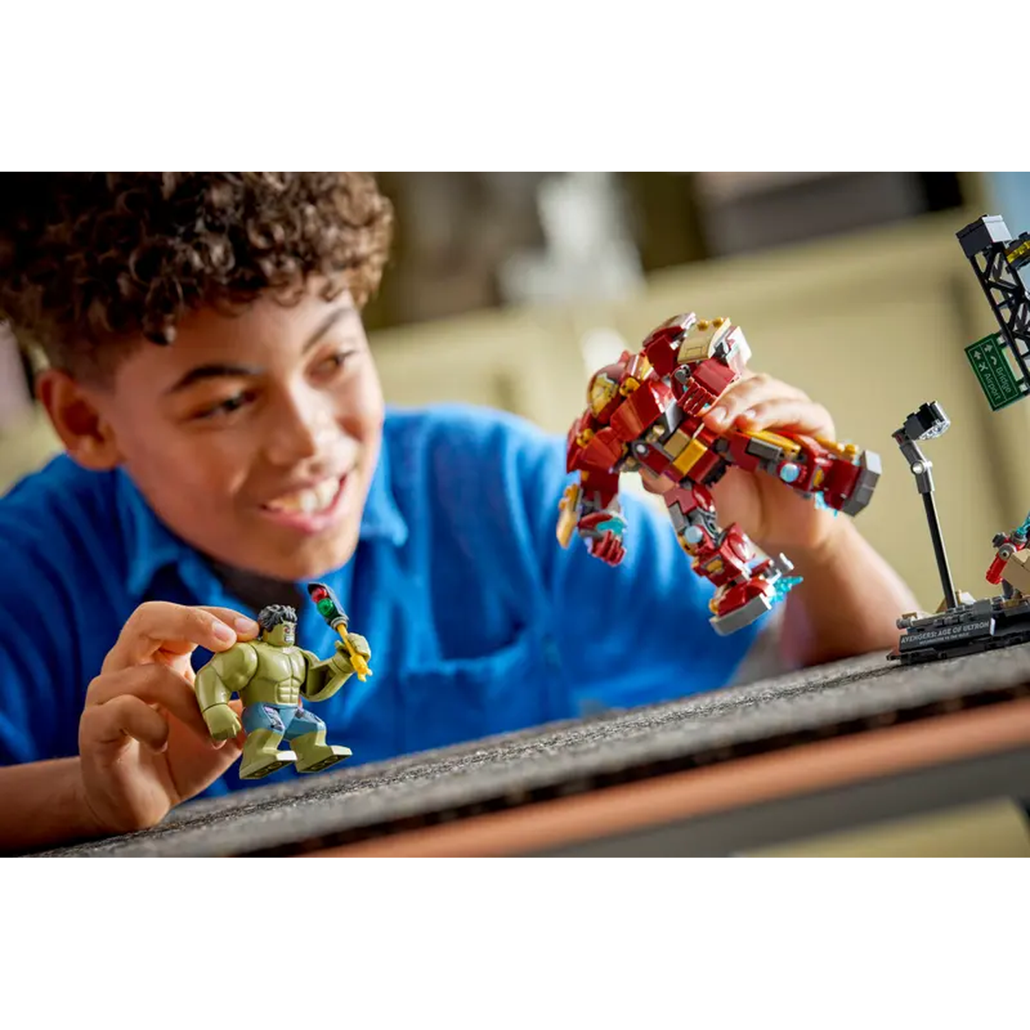 A smiling boy plays with Lego figures of Hulk and Iron Man. The setting includes a bridge and airport sign in the background.