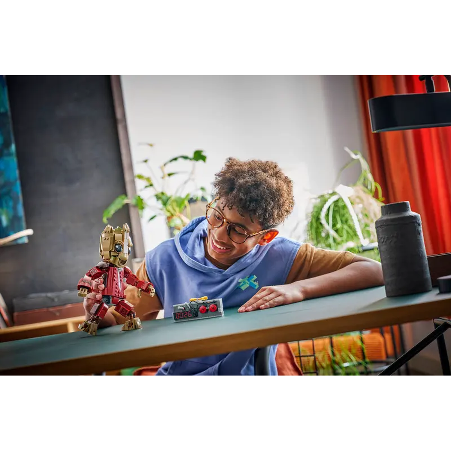 A smiling boy with glasses looks at a Lego Groot figure. A small timer sits next to it. In the background are plants and a dark vase.