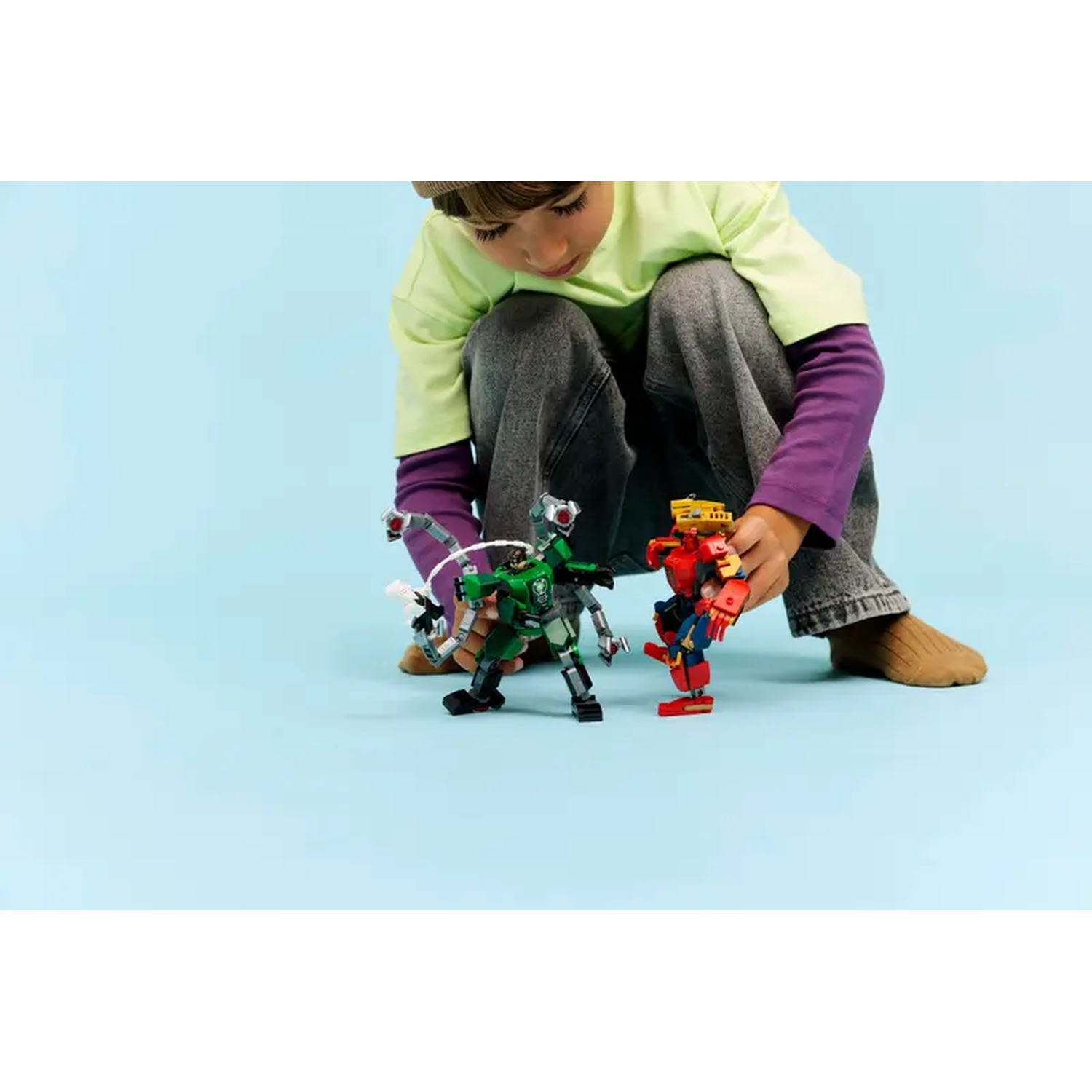 A child playing with toy robots. The child is wearing a green shirt and gray pants. Robots are green, red and yellow.