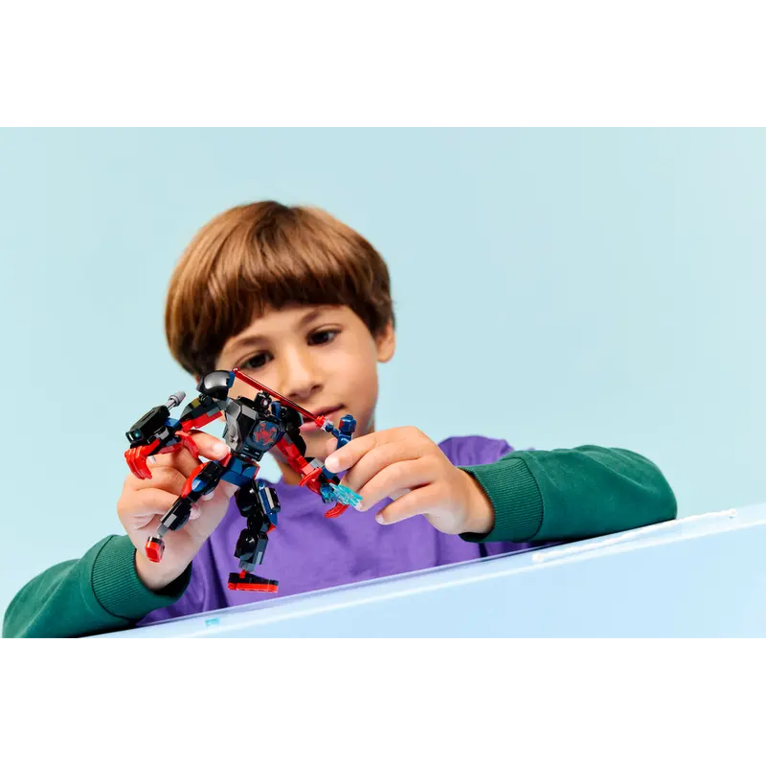 A boy holds two toy robots, one black and red, the other blue, against a light blue backdrop. He wears a purple shirt.