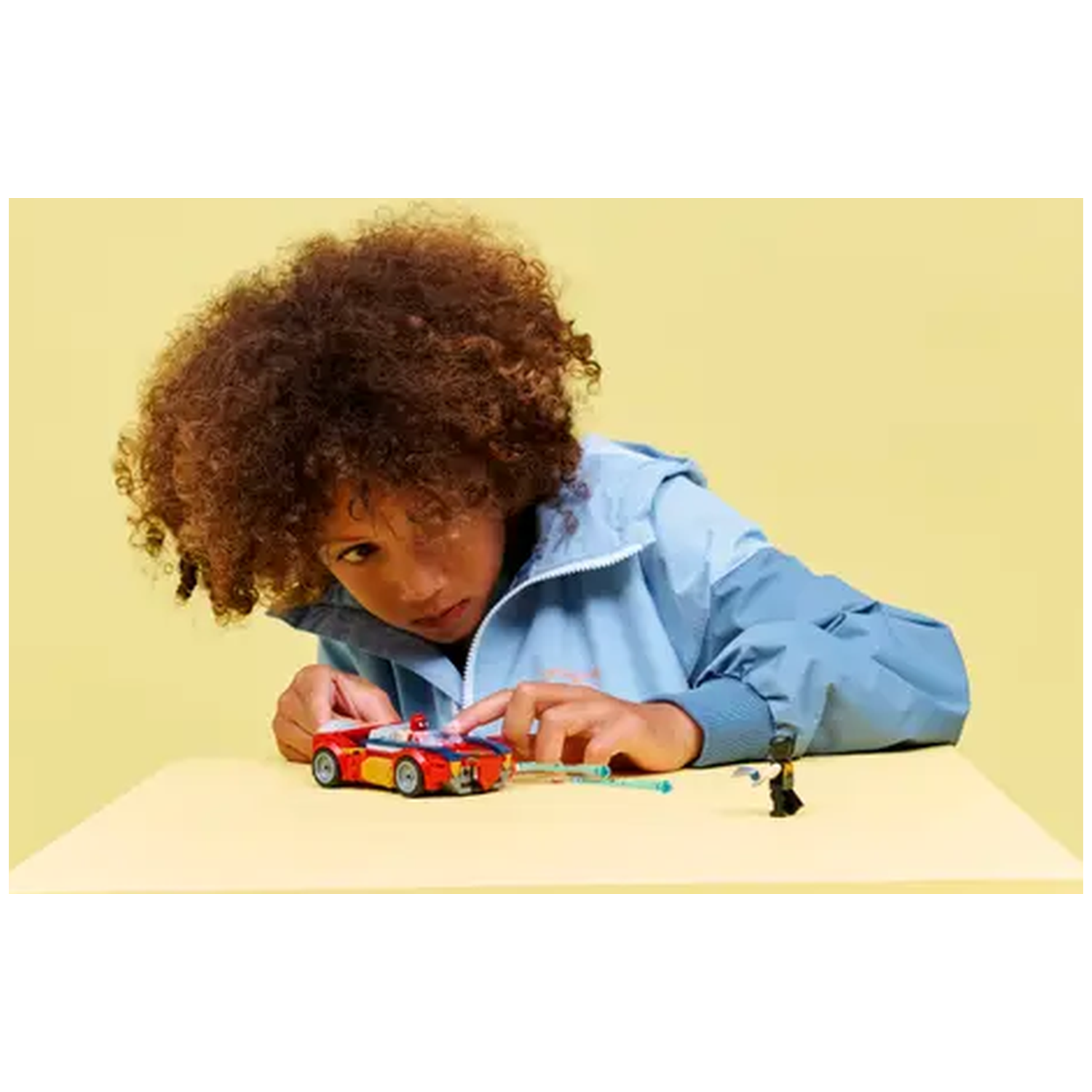 A child in a blue jacket looks at a red and yellow toy car on a yellow table. A black Lego figure stands nearby.