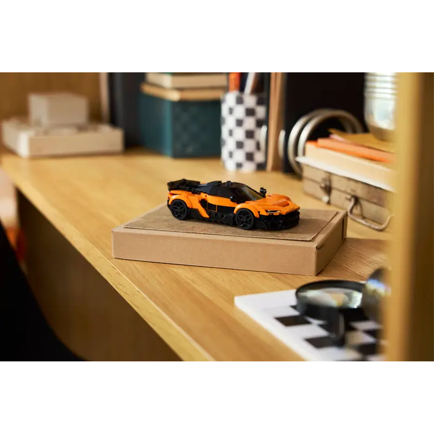 Een oranje en zwarte speelgoedauto staat op een kartonnen doos op een houten bureau. Boeken, pennen en een vergrootglas zijn zichtbaar.