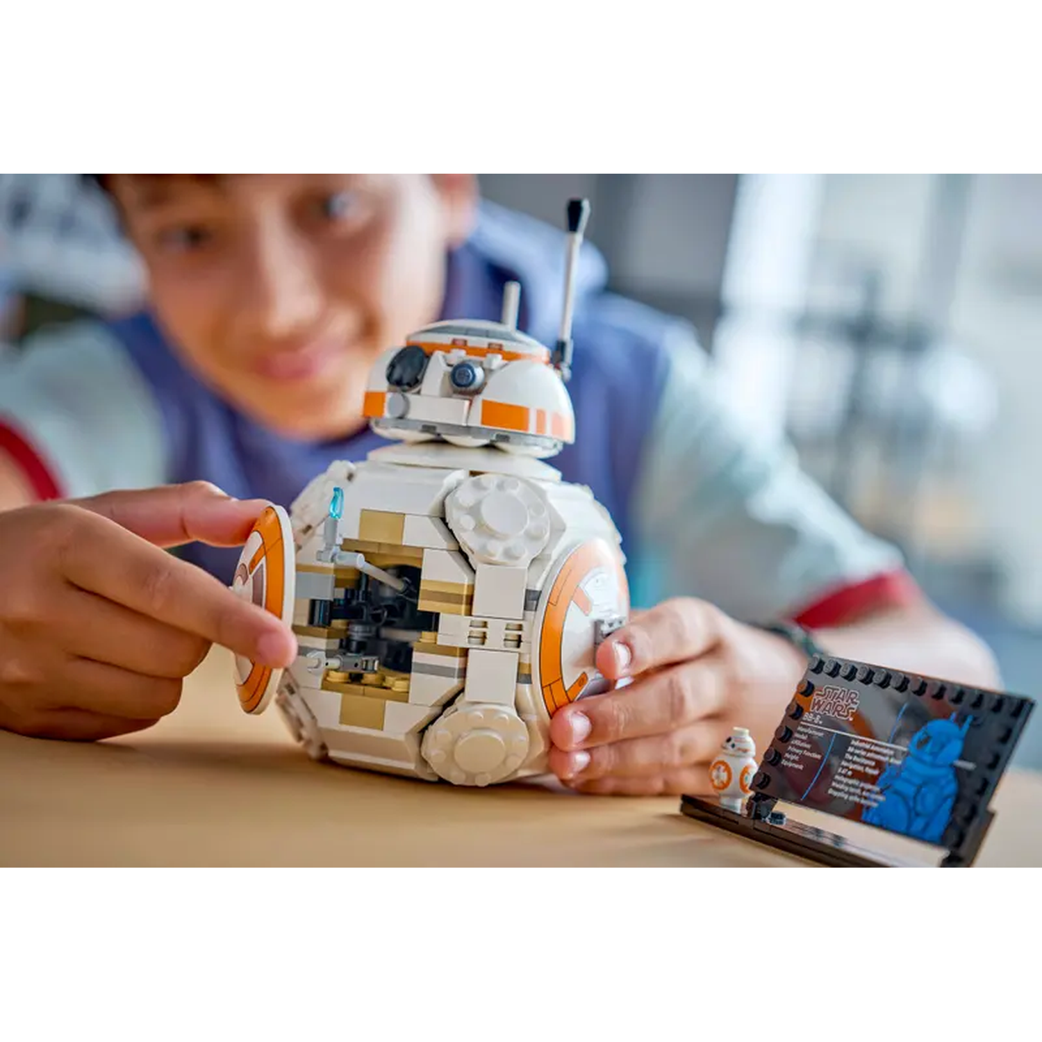 Een jongen monteert een witte en oranje BB-8 Lego-set. Een kleinere BB-8 figuur en een Star Wars info kaart liggen op de tafel.