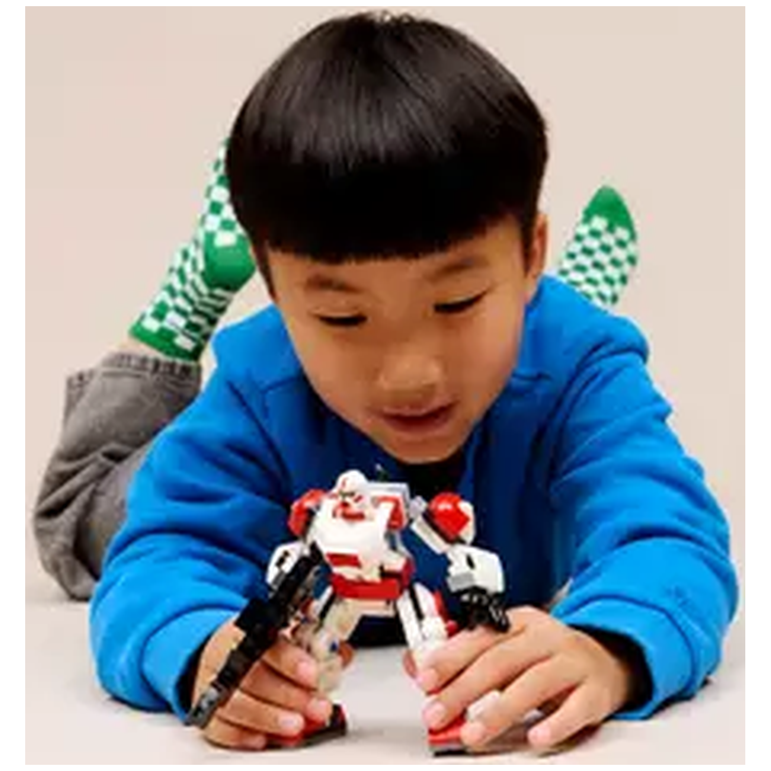 Een jongen in een blauw shirt speelt met een robotspeeltje. Hij ligt. Groene geruite sokken zijn zichtbaar op de achtergrond.