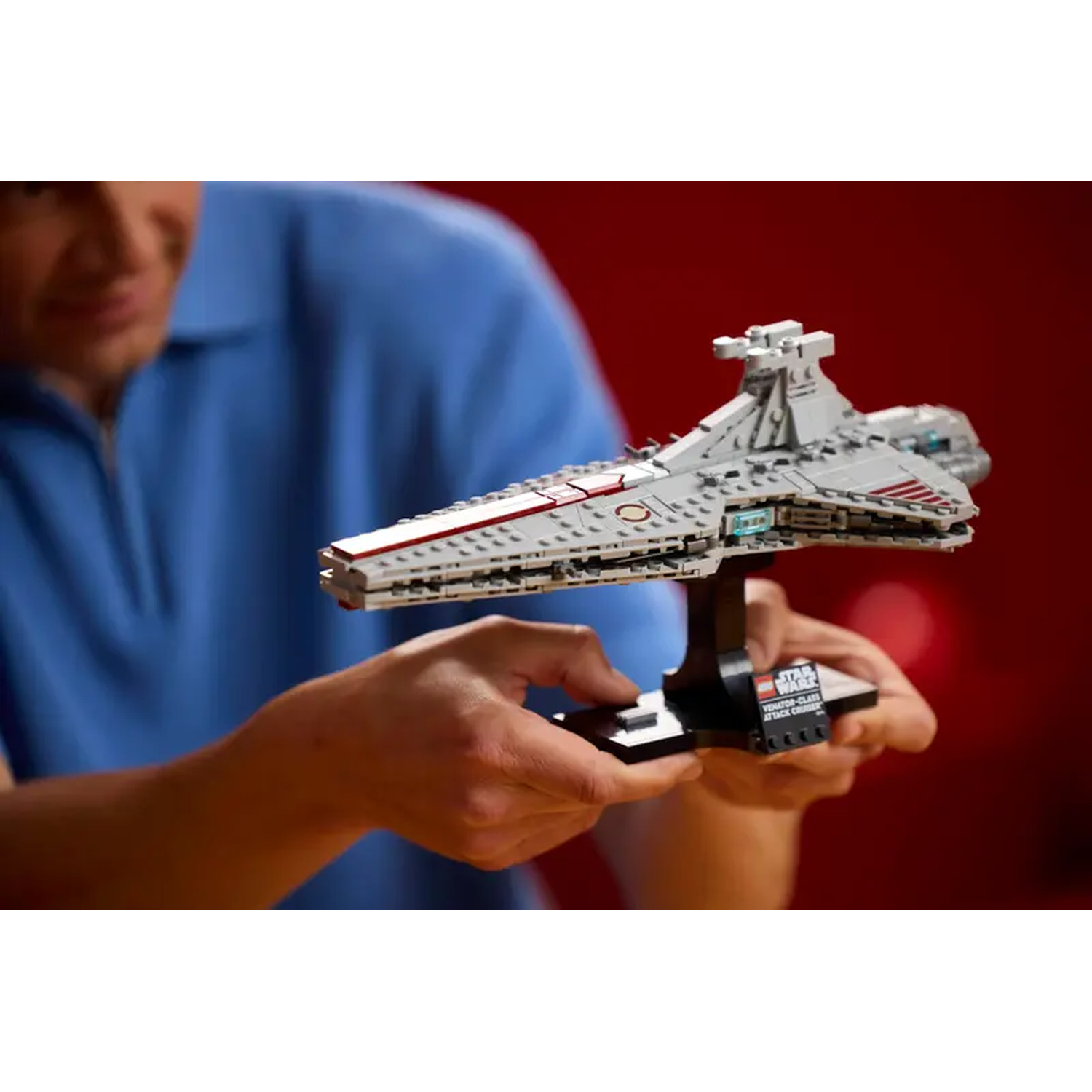 Een persoon houdt een Star Wars Venator-class Attack Cruiser LEGO-set vast. Het model is grijs, met rode accenten, en staat op een zwarte standaard.