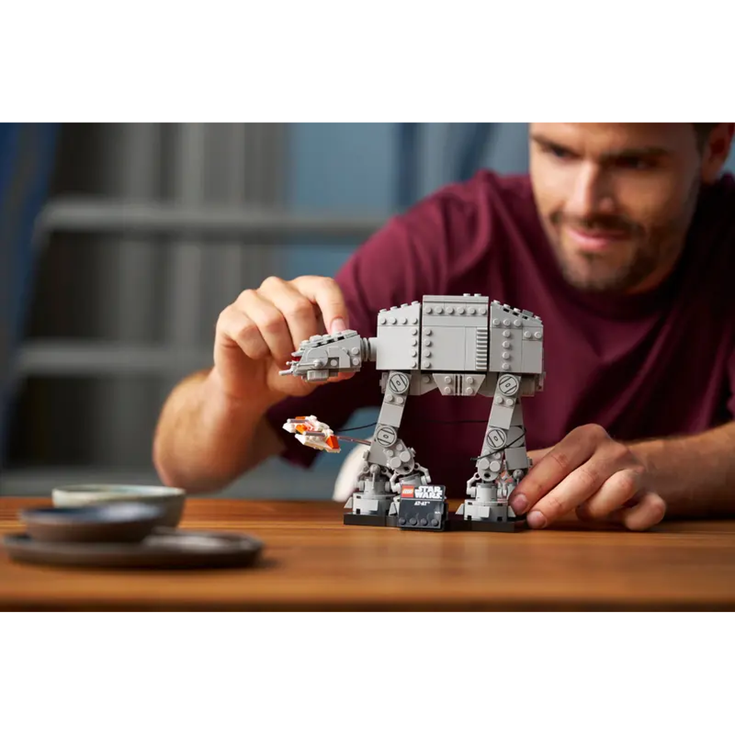 Een man die een Lego AT-AT-model in elkaar zet op een houten tafel. Het model is grijs en de man draagt een bordeauxrode shirt. Andere objecten zijn zichtbaar.