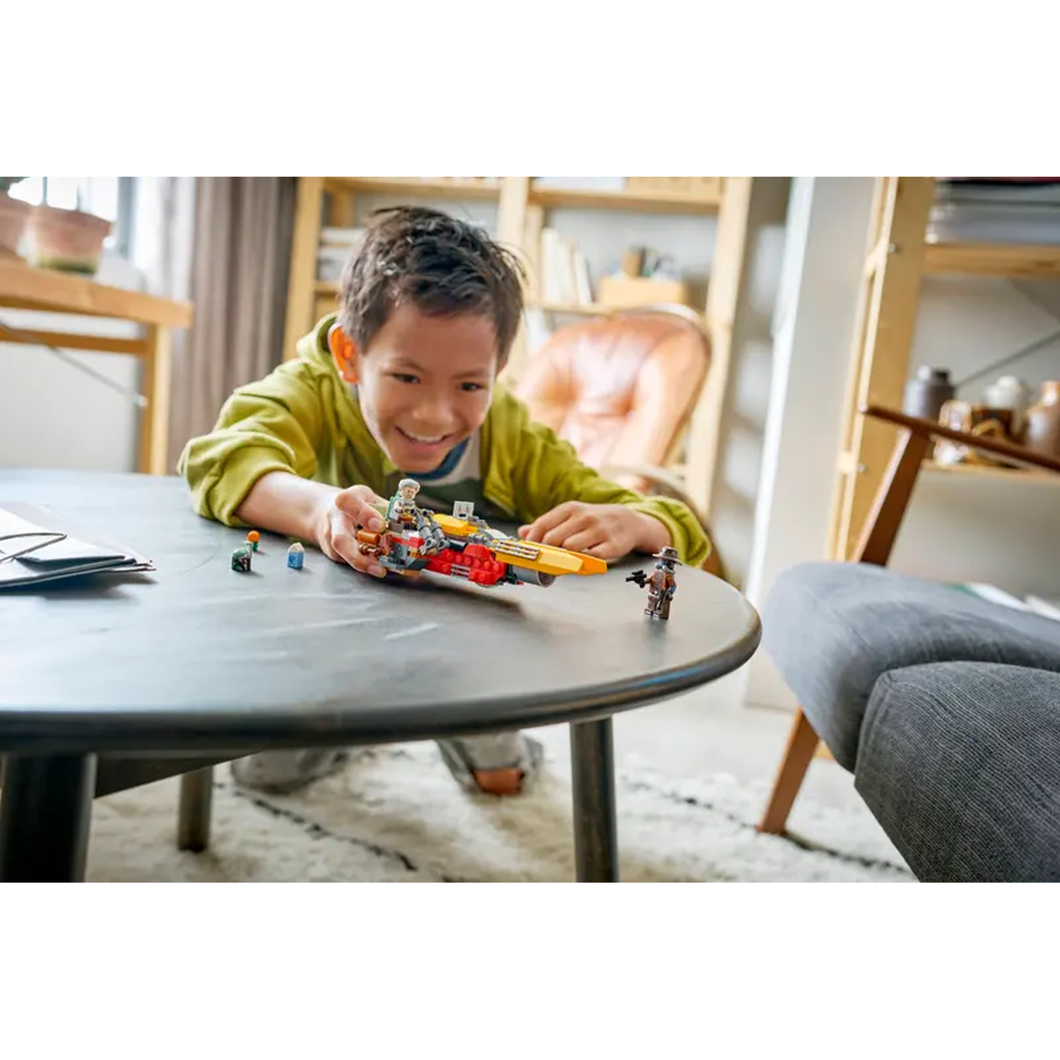 A smiling boy plays with Lego on a round table. Lego figures and a vehicle are in front of him. A couch and bookshelf are in the background.
