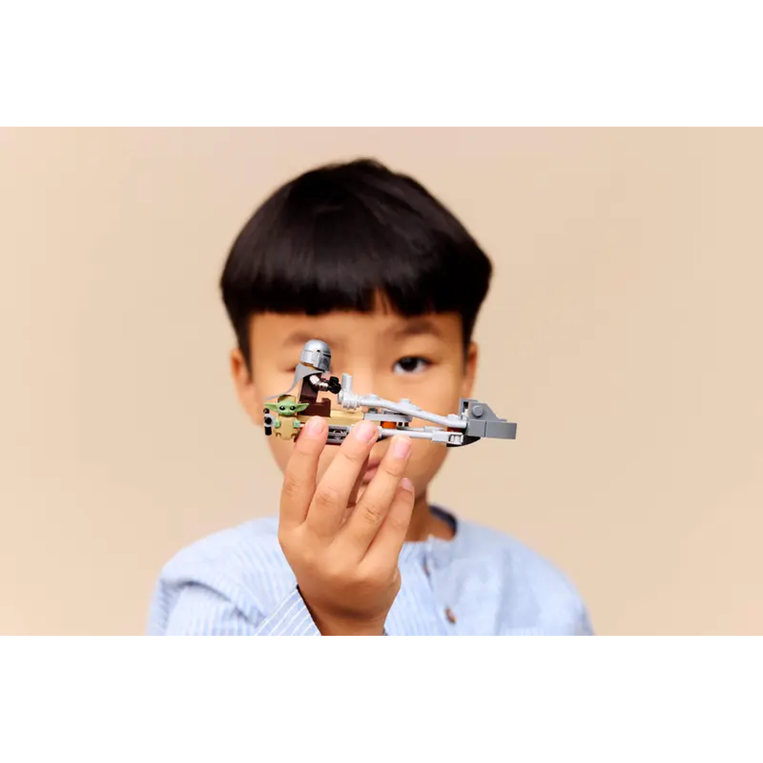 A child holds a Lego Star Wars speeder. The child has dark hair and is wearing a blue shirt. Background is a beige wall.