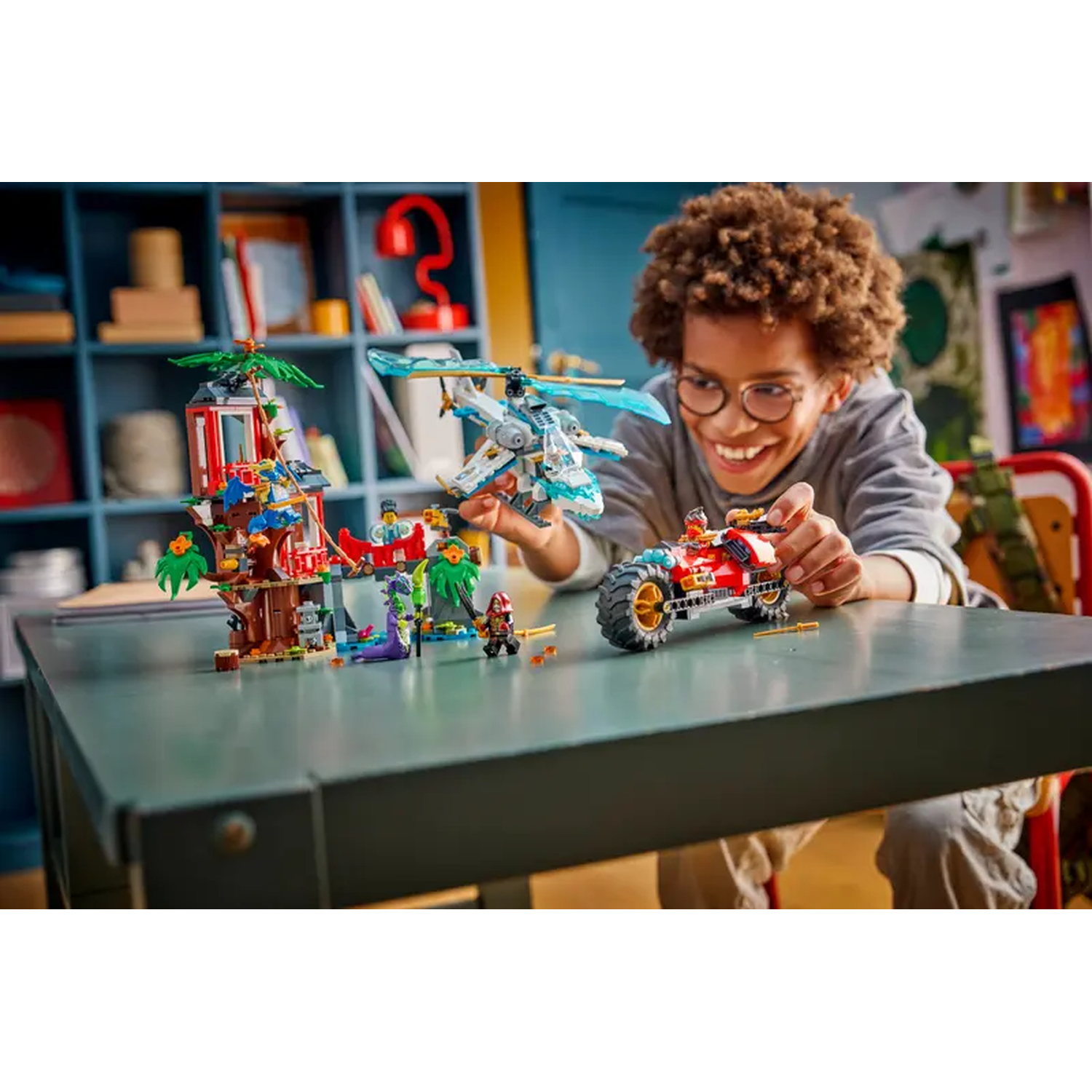A smiling boy with glasses plays with Lego sets, including a red motorcycle, a helicopter, and a multi-story building.