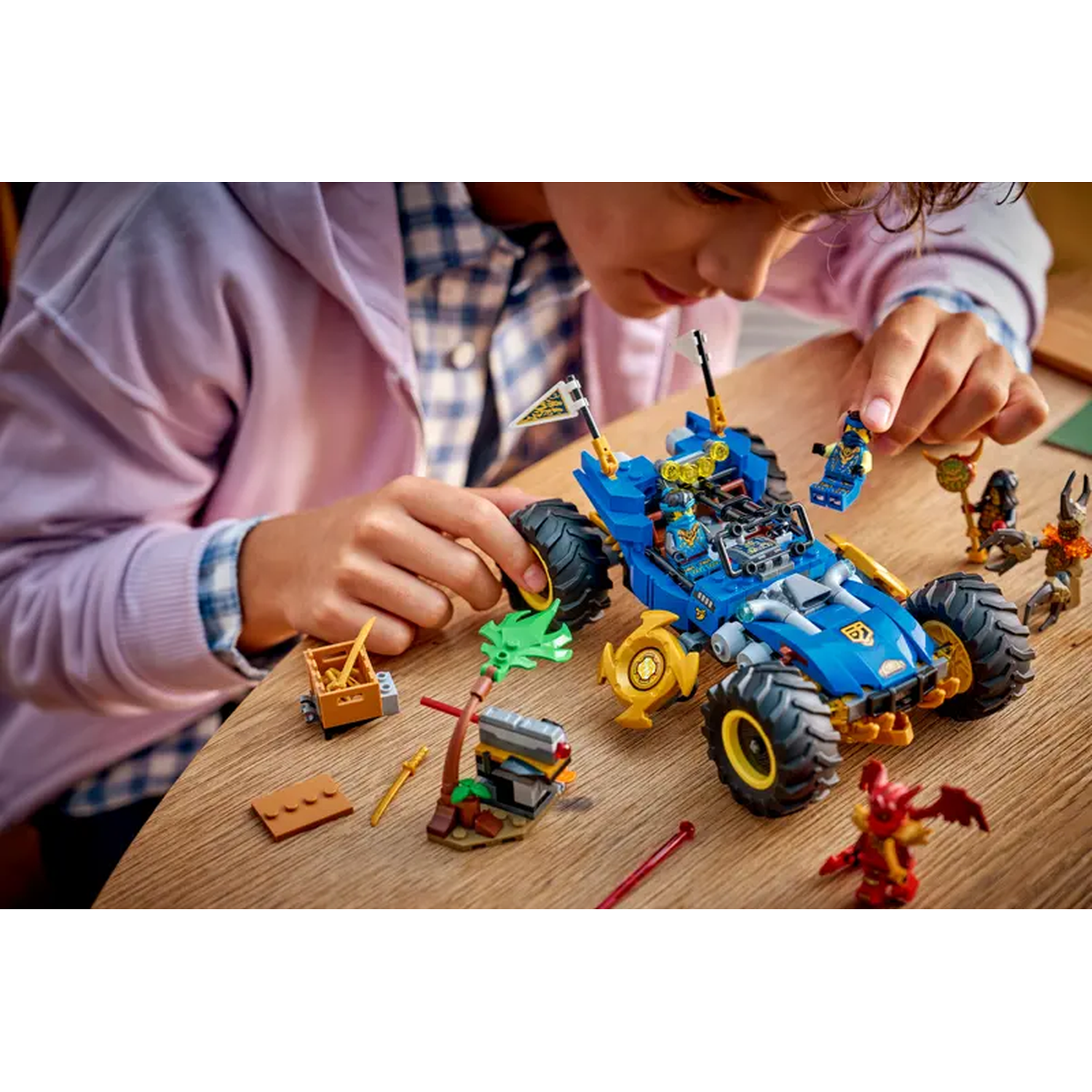 A person assembling a blue Lego car with yellow accents, surrounded by other Lego figures and structures on a wooden surface.