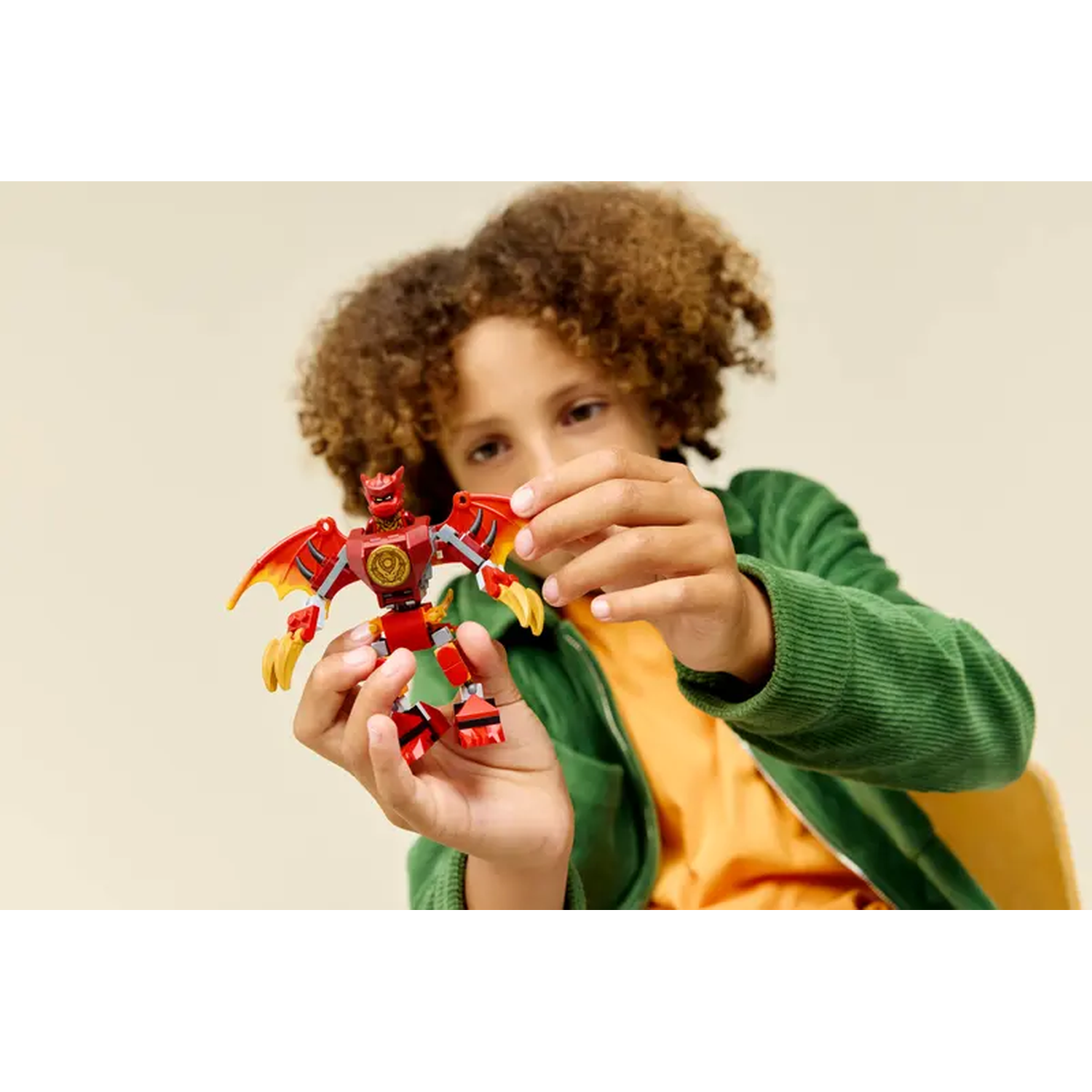 A child holds a red and yellow Lego dragon toy. The child wears a green jacket and orange shirt. Beige background.