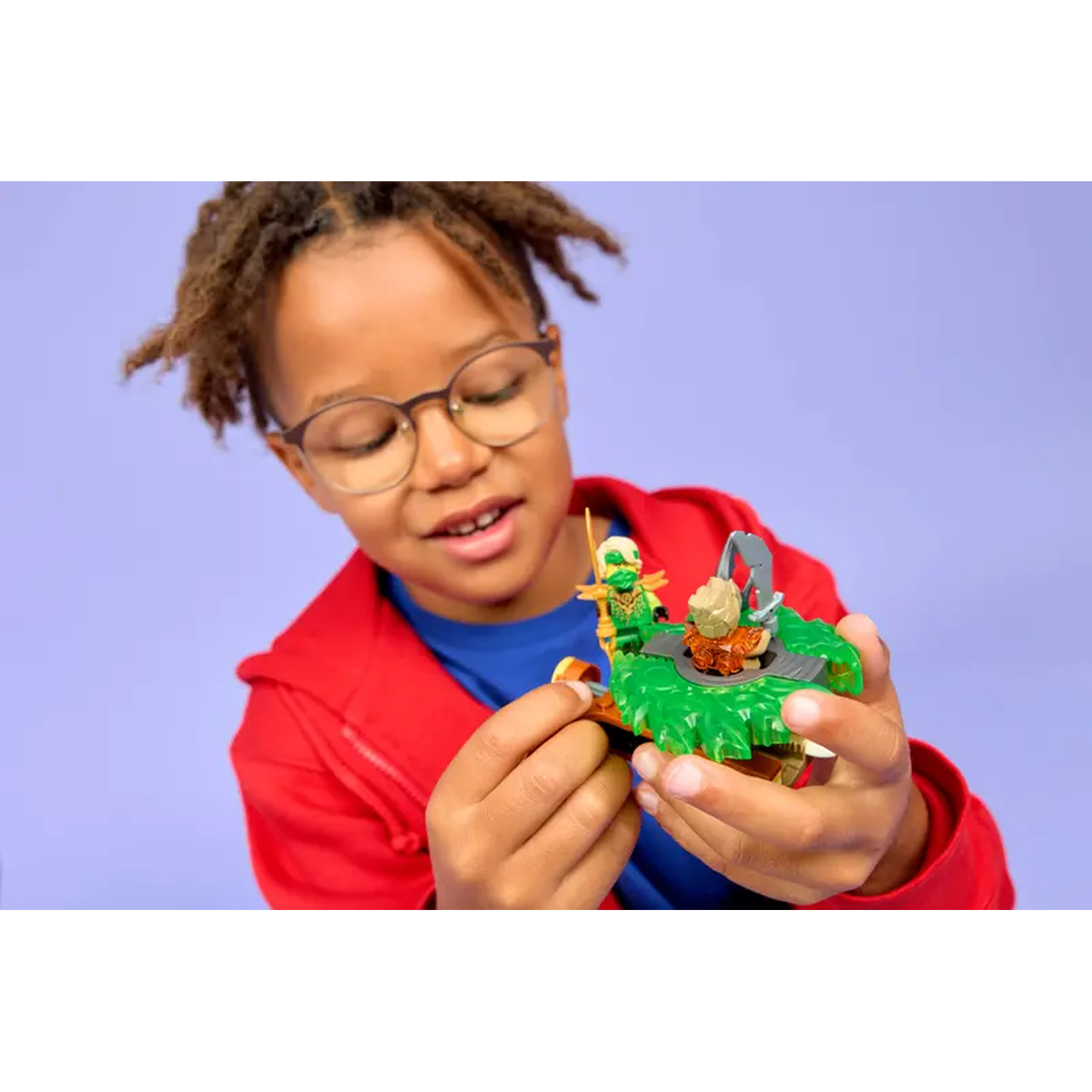 A young boy with glasses holds a Lego set. The set features a green base with a character and other Lego pieces.