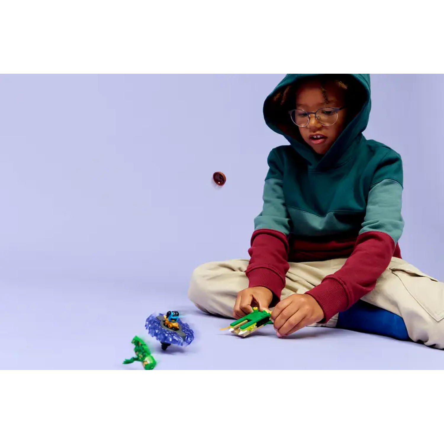 A child in a hoodie and glasses sits playing with toys. A small brown object is floating in the air. Toys are on the floor.
