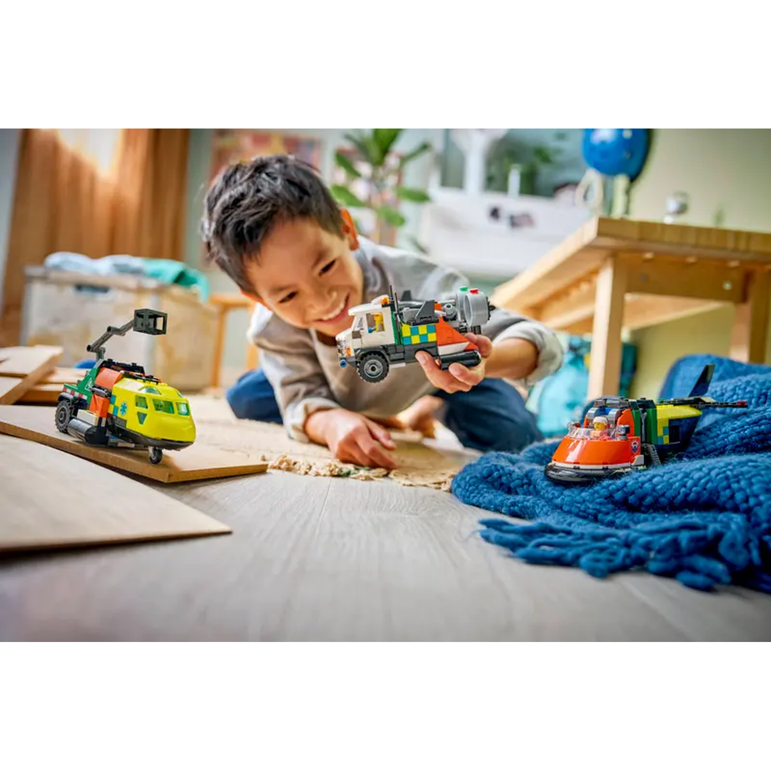 A boy smiles, holding a white and red toy truck. Two other toy vehicles are nearby. The scene is indoors.