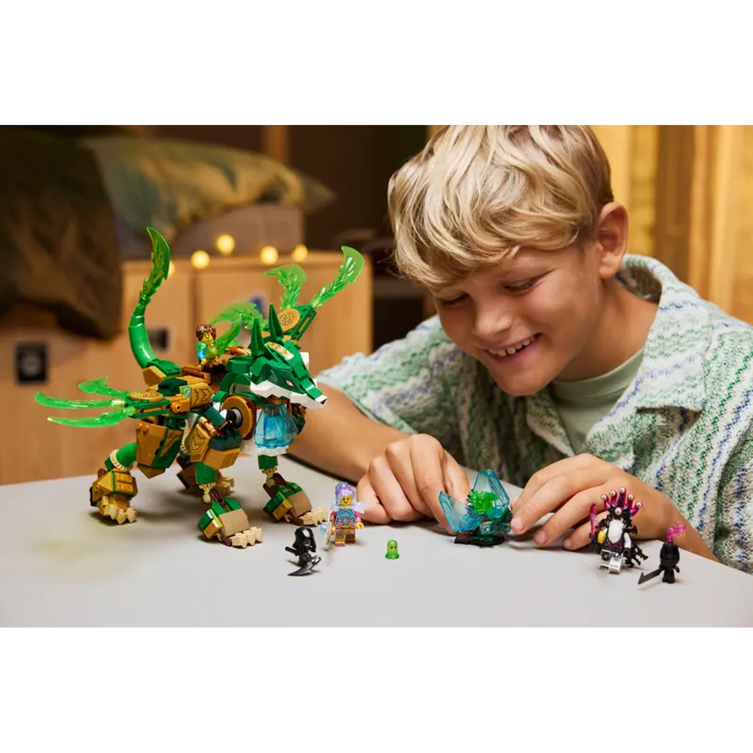 Een jongen lacht, spelend met een Lego-draak. De draak is groen en goud. Kleine figuren staan op de tafel. Er is een blauwe kristal.